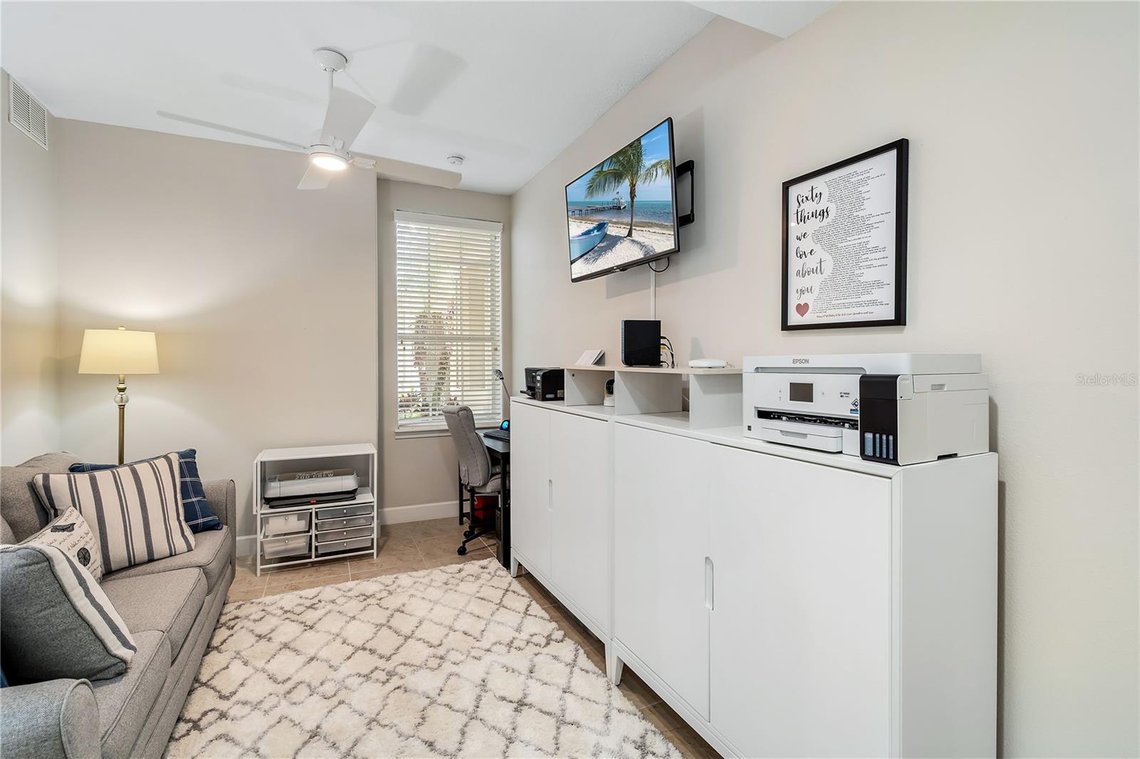 Furnished home office with a gray sofa, white cabinets, printer, and wall-mounted television.
