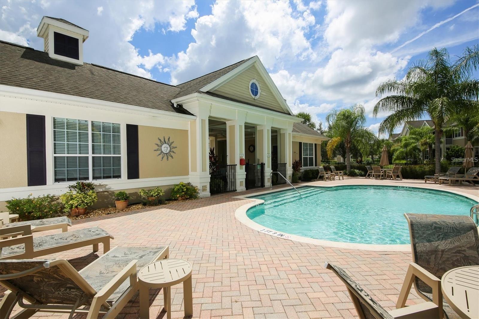 Community pool beside a columned clubhouse, with brick paving, lounge chairs, and palm trees.