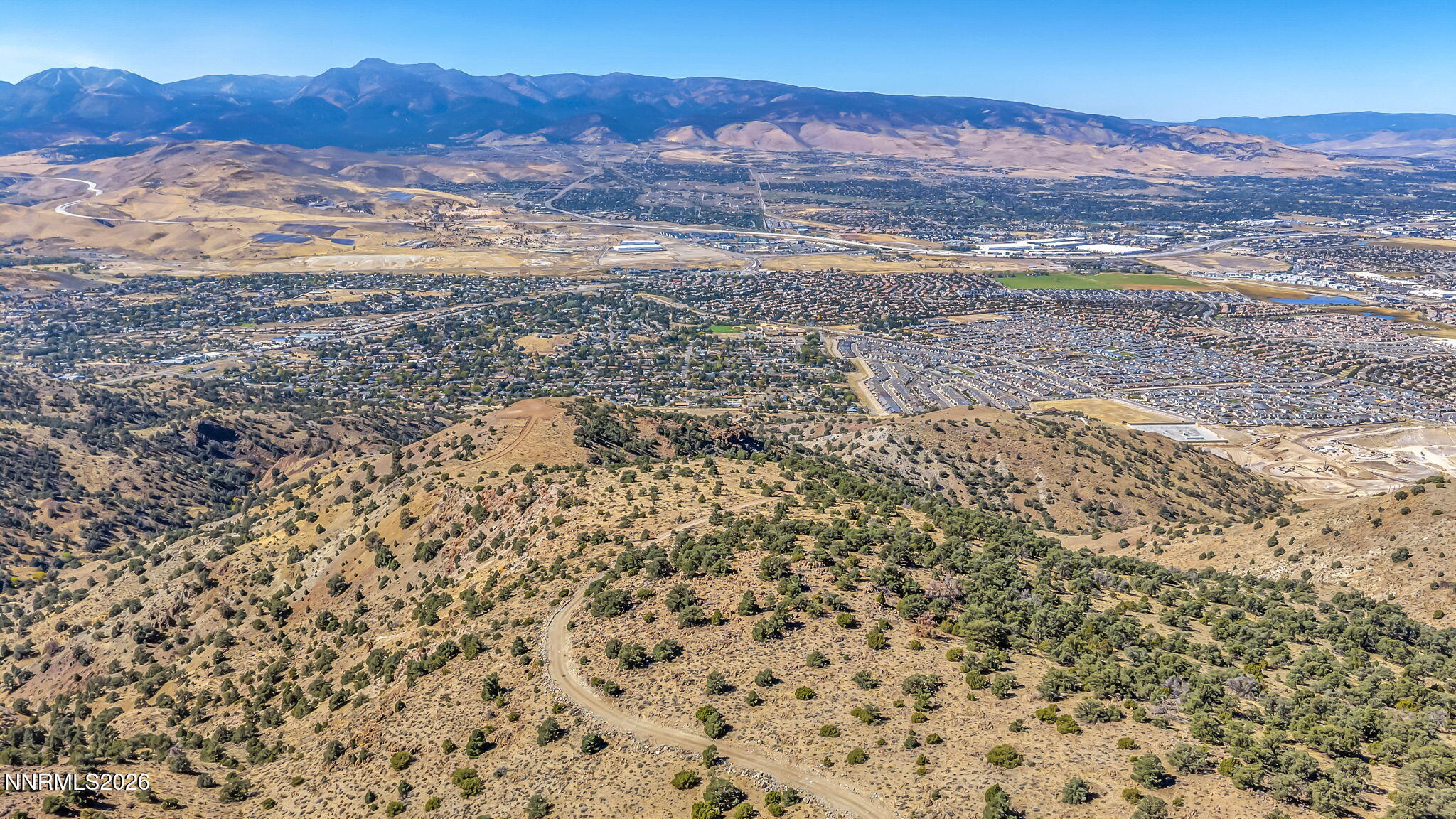 Aerial view across a Reno valley, with a winding dirt road, sprawling neighborhoods, and distant mountains.