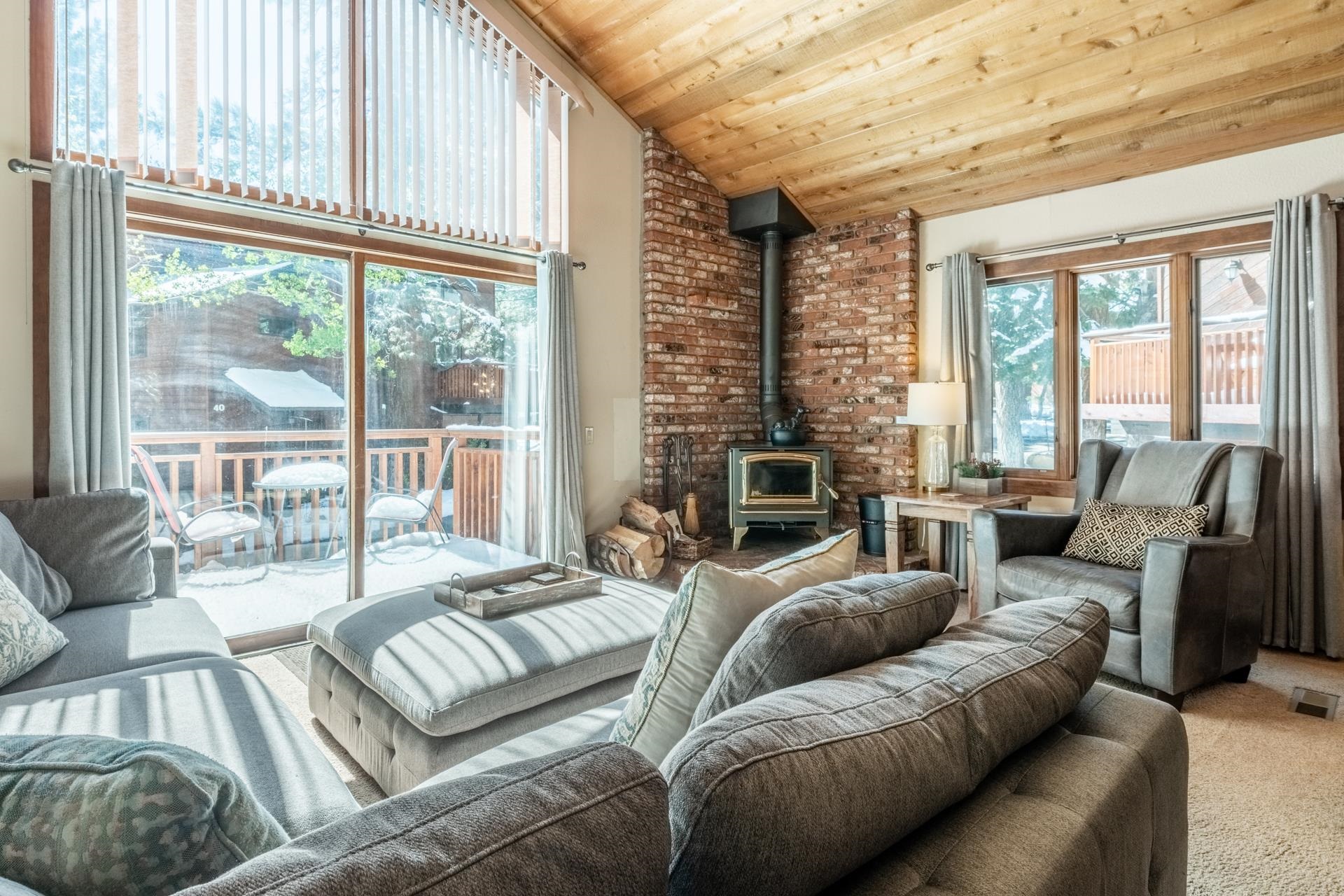 Furnished living room with vaulted wood ceiling, brick fireplace, and snowy deck views.
