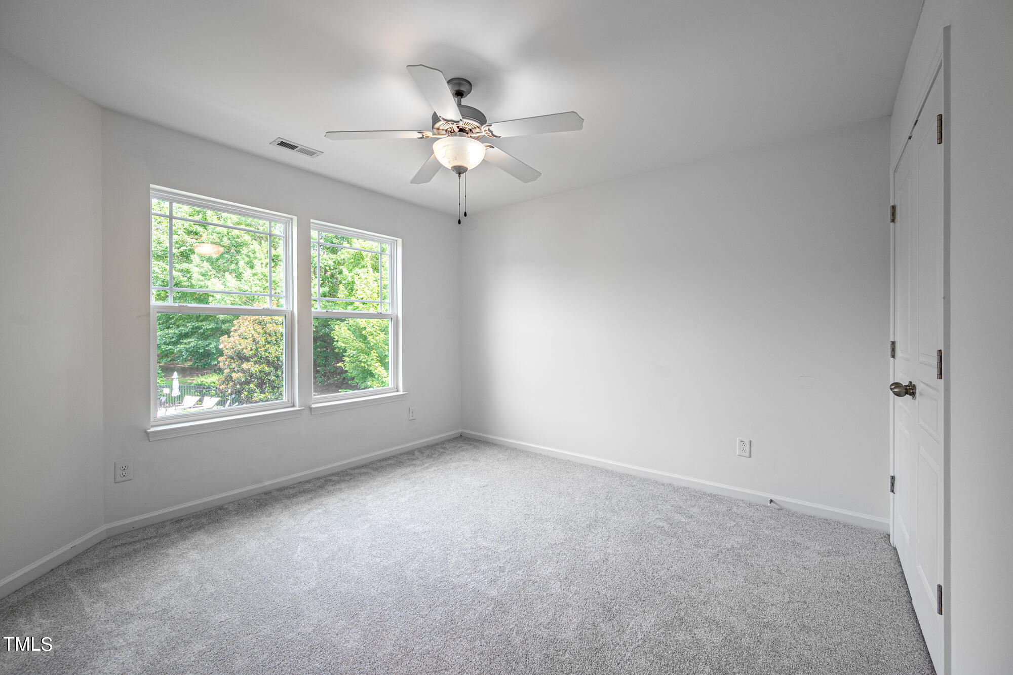 Empty bedroom with gray carpet, two windows overlooking greenery, and a ceiling fan with light.
