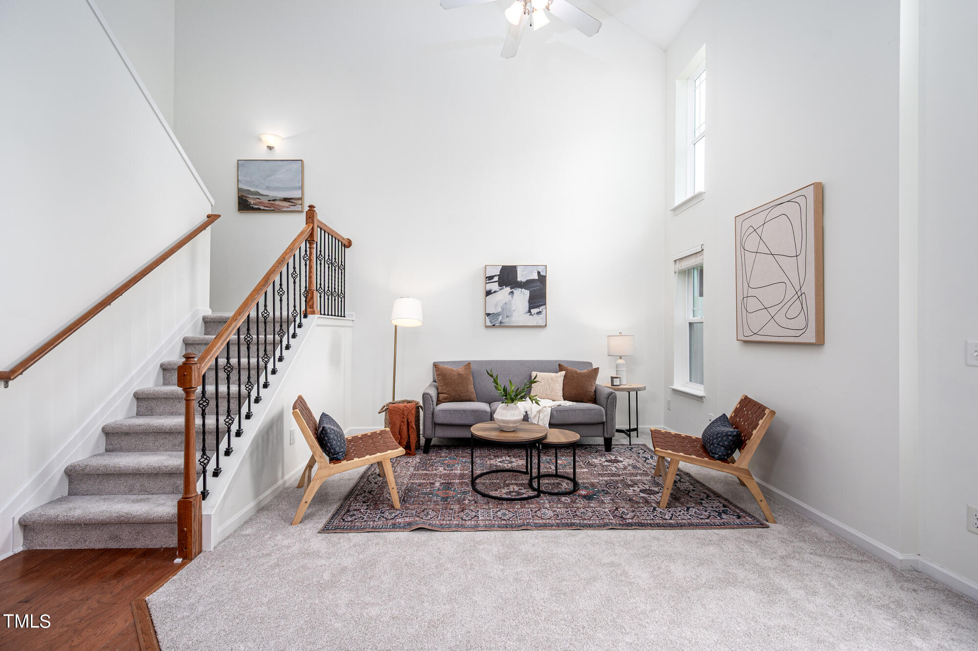 Staged living room with vaulted ceiling, carpeted staircase, gray sofa, and tall windows.