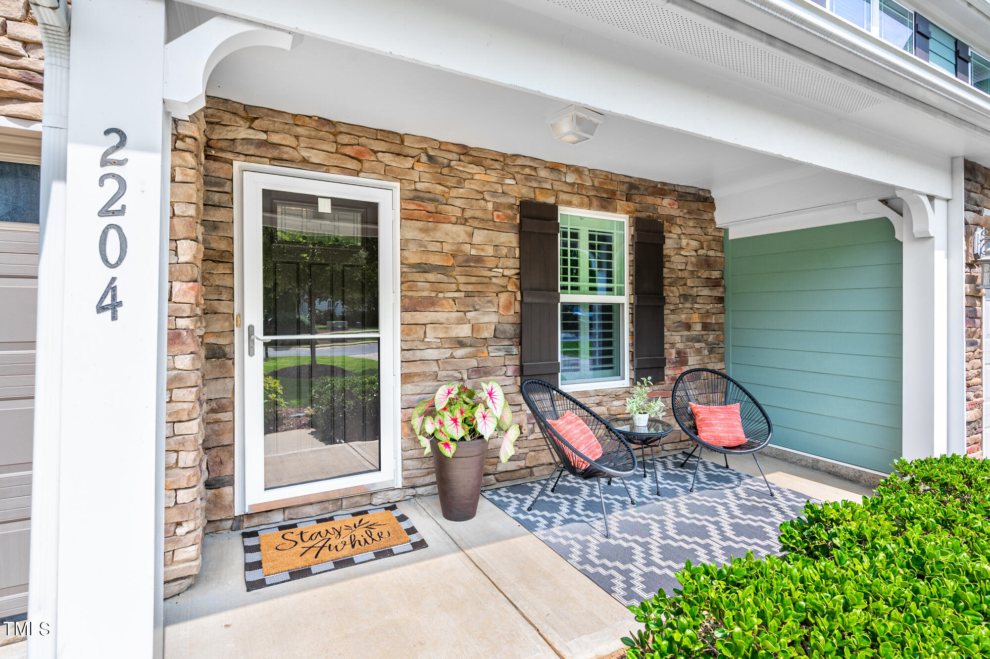 Covered stone-front porch with two woven chairs, coral cushions, a patterned rug, and a potted plant.
