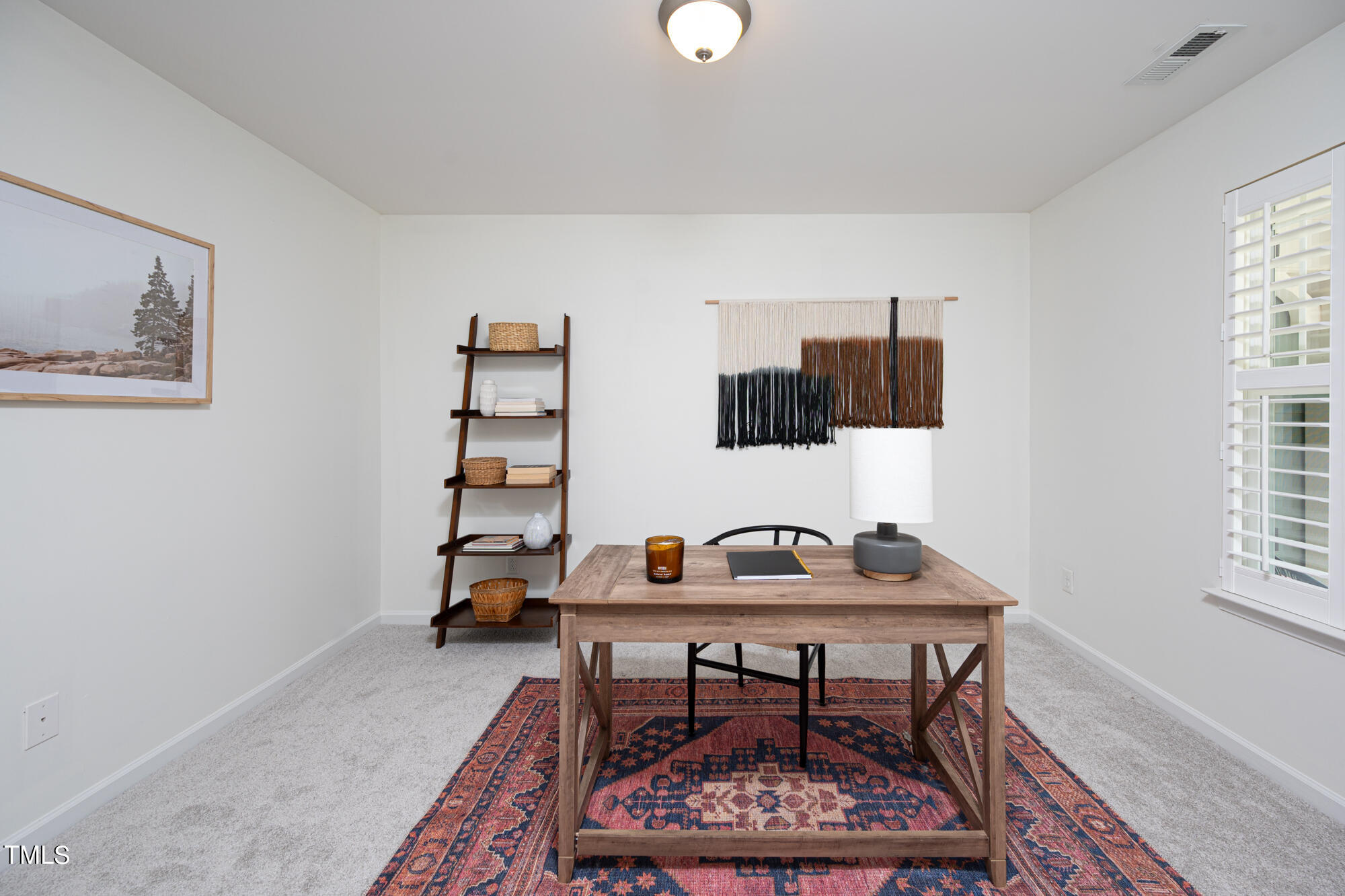 Staged home office with a wooden desk, patterned rug, ladder shelf, and shuttered window.