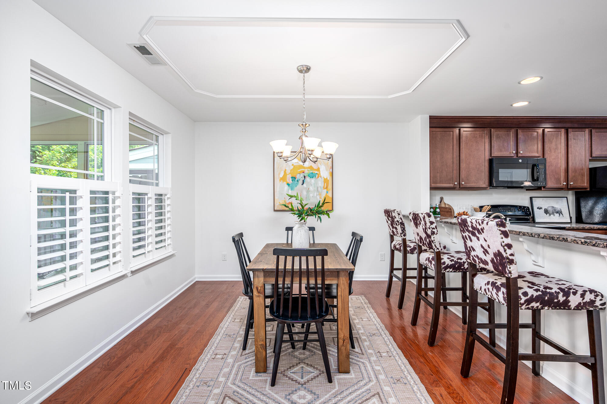 Dining area with a wooden table, plantation-shuttered windows, and a kitchen breakfast bar with cow-print stools.