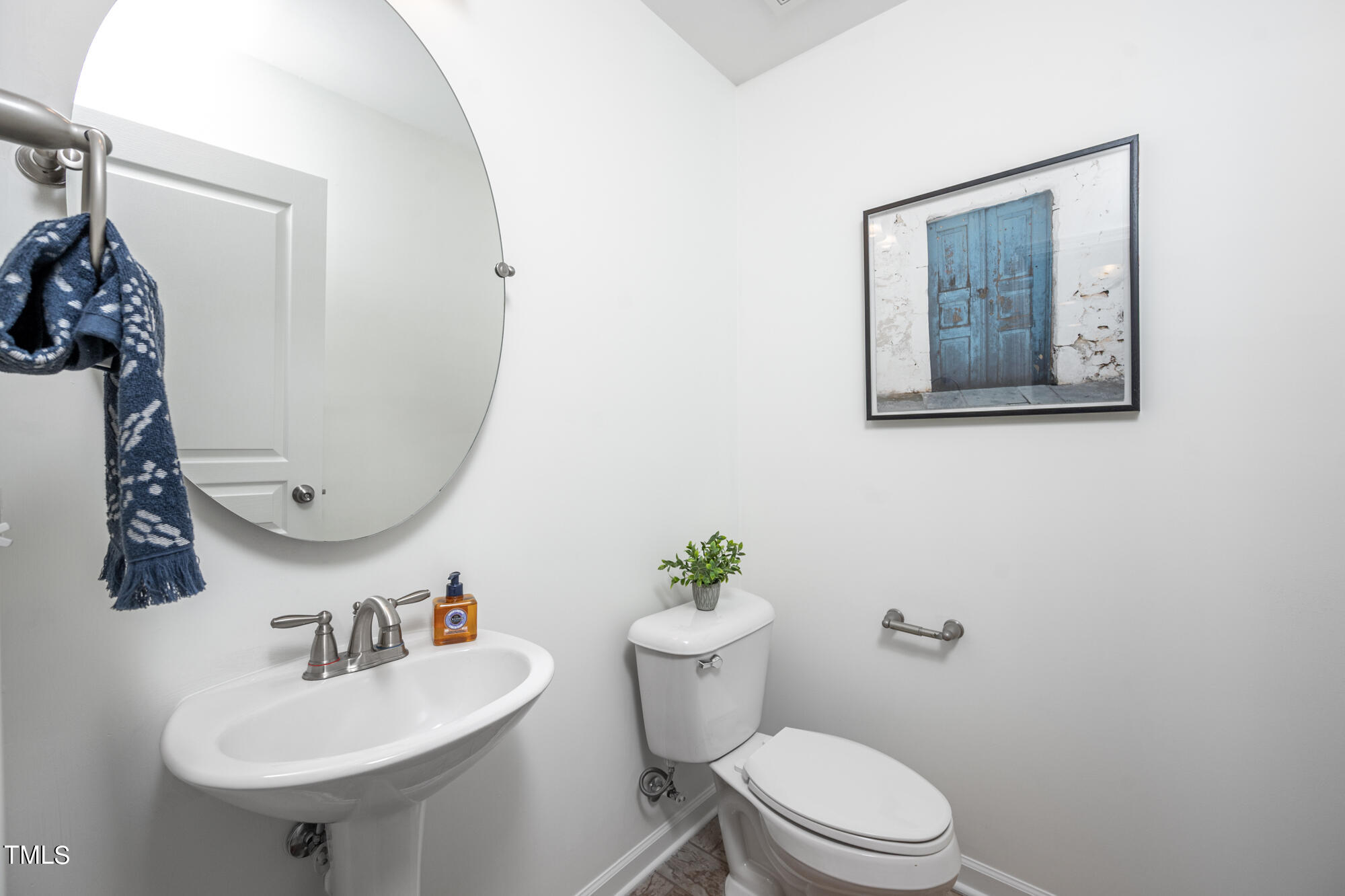 Half bathroom with a pedestal sink, round mirror, toilet, blue towel, and framed blue-door artwork.