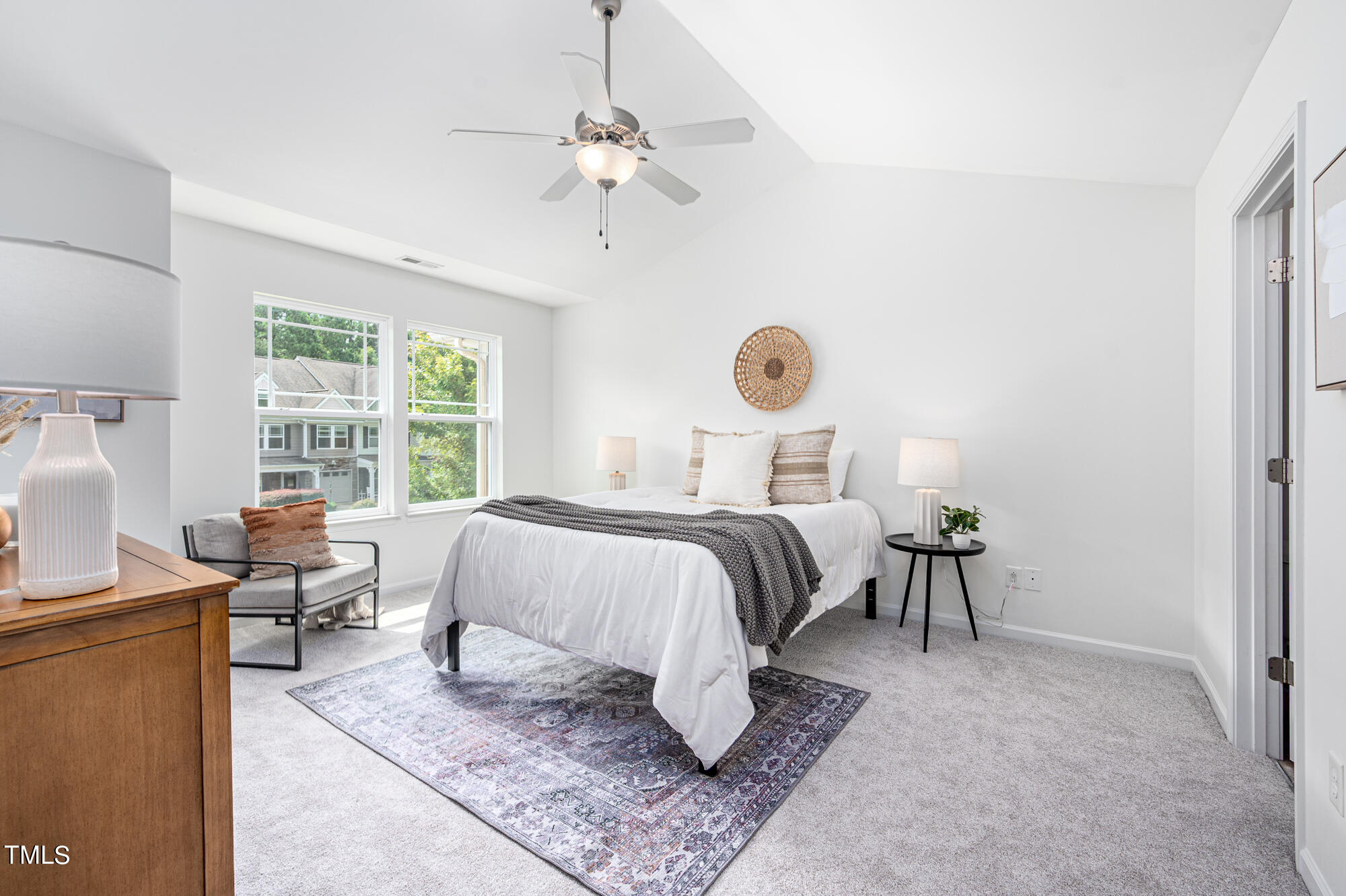 Bright bedroom with a white bed, patterned rug, ceiling fan, and windows overlooking trees.