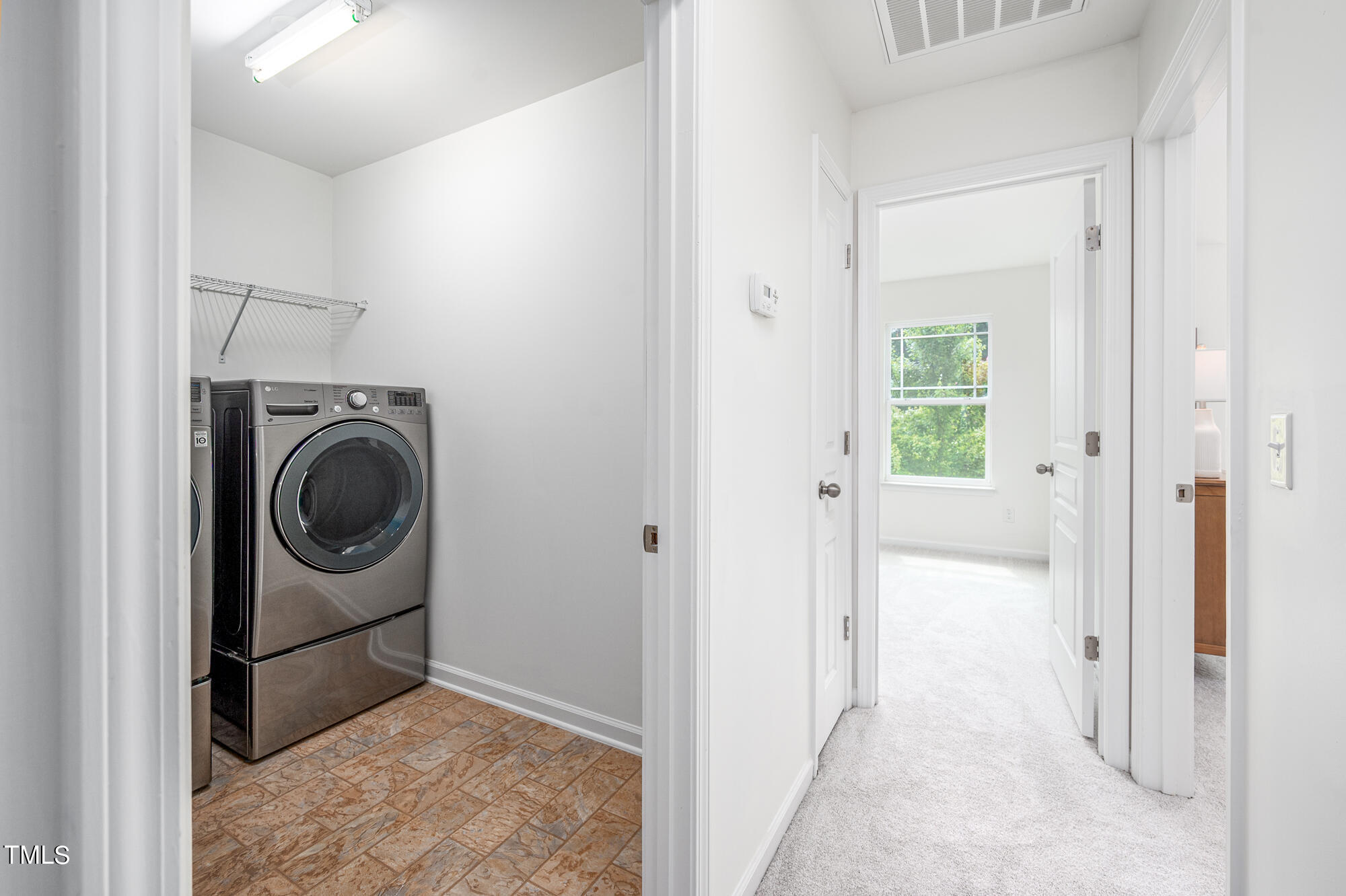 Laundry room with stacked washer and dryer beside a carpeted hallway leading to a bright room.