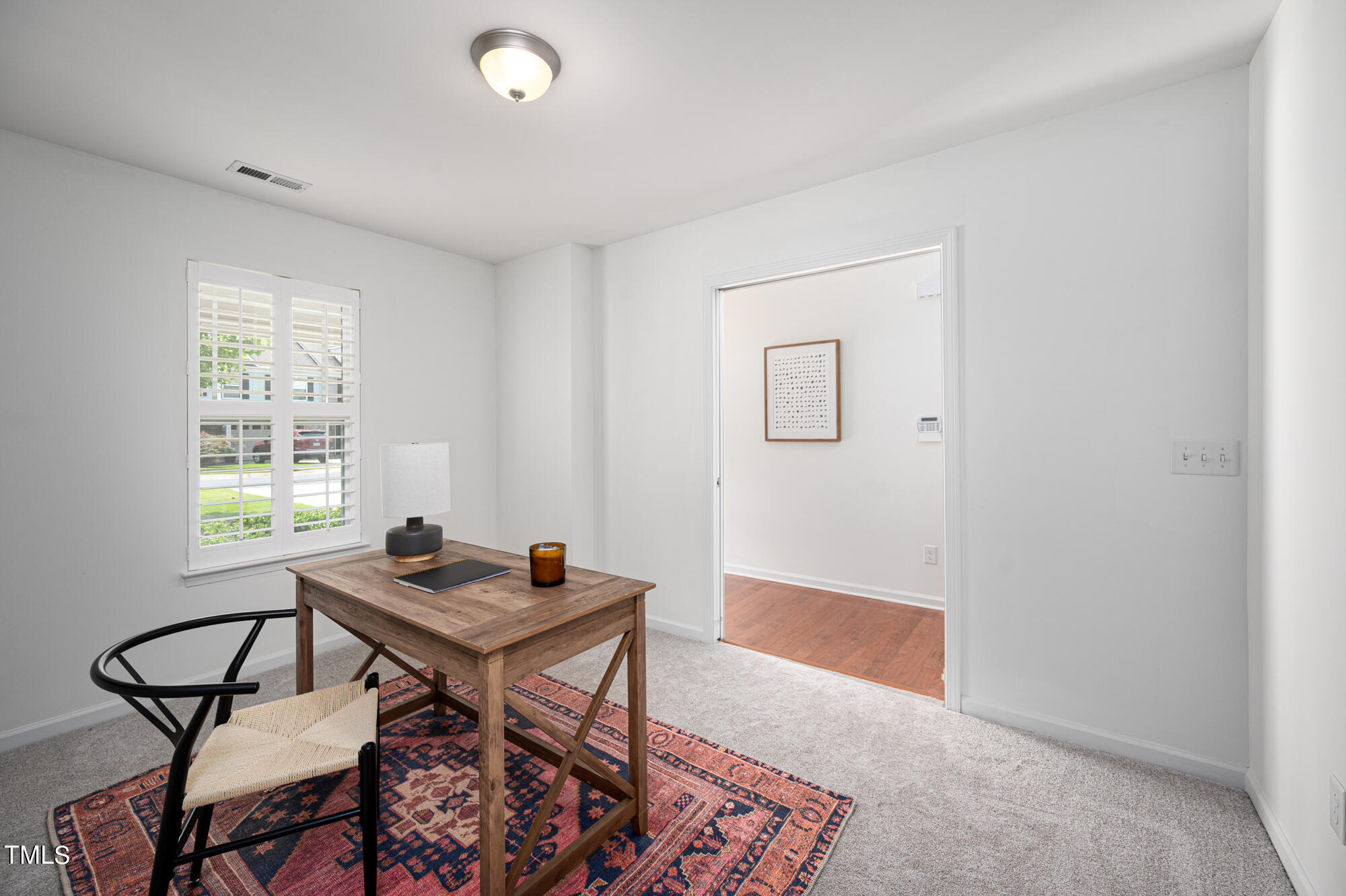 Staged home office with a wooden desk, patterned rug, and plantation-shuttered window.