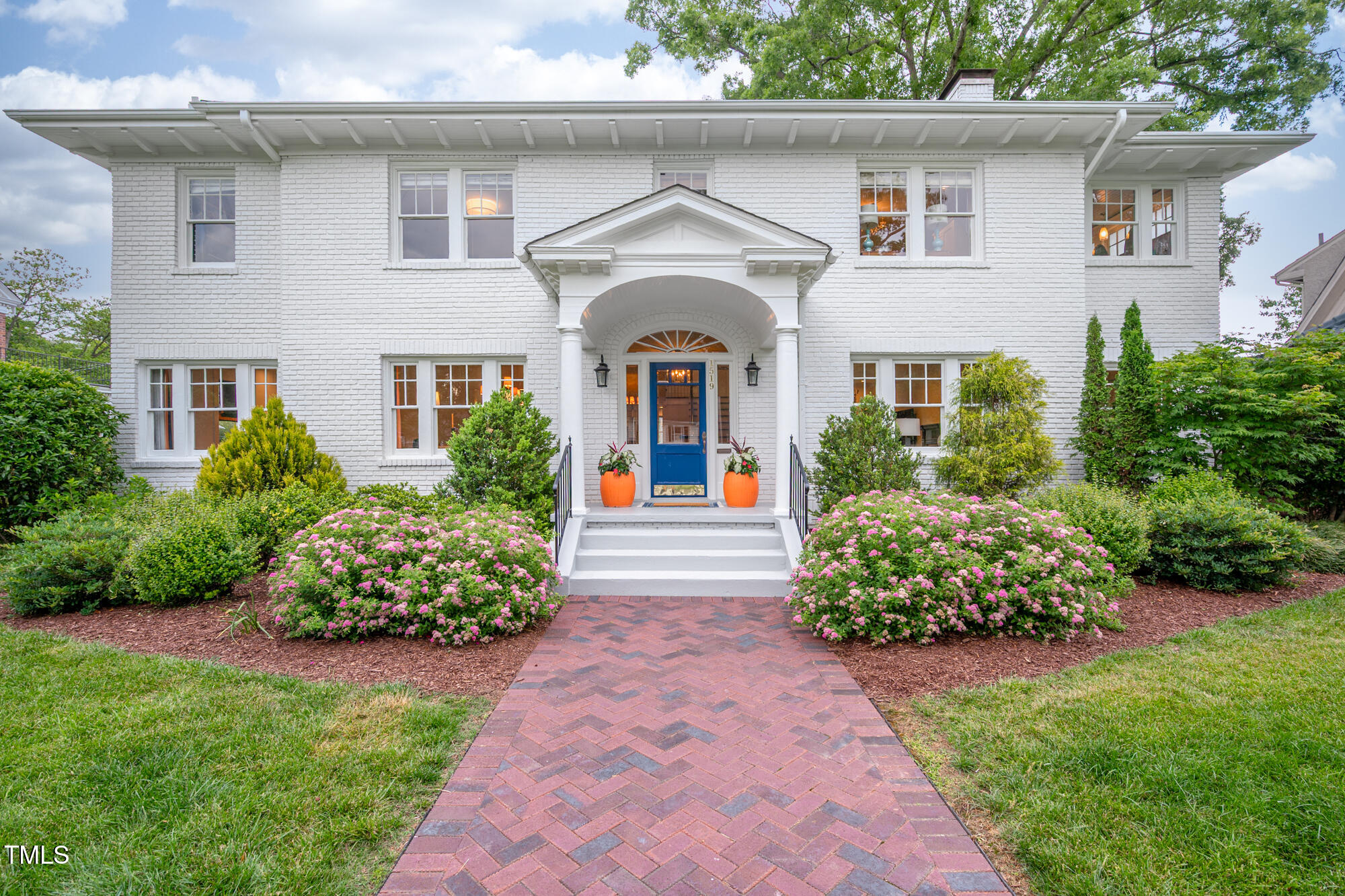 White brick house with a blue front door, columned entry, and pink flowering shrubs along the brick walkway.
