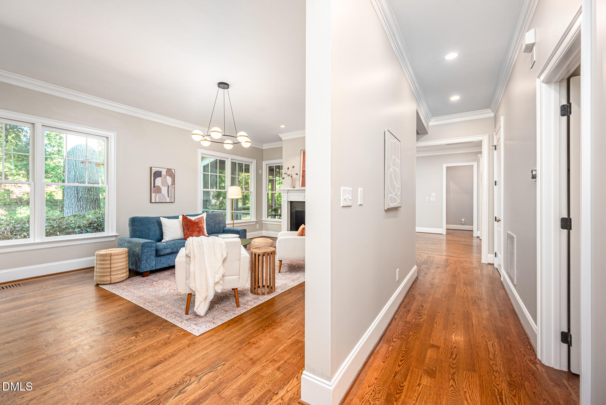 Staged living room beside a hardwood hallway, with large windows, a blue sofa, and a fireplace.