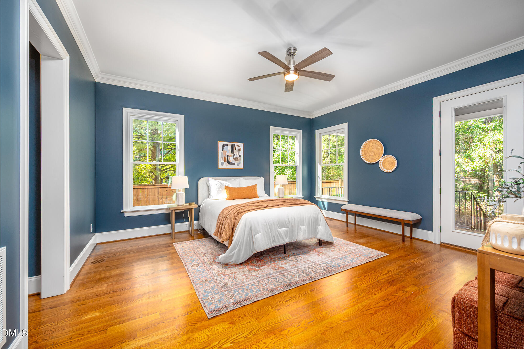 Staged bedroom with deep blue walls, a white bed, hardwood floors, and tall windows overlooking greenery.