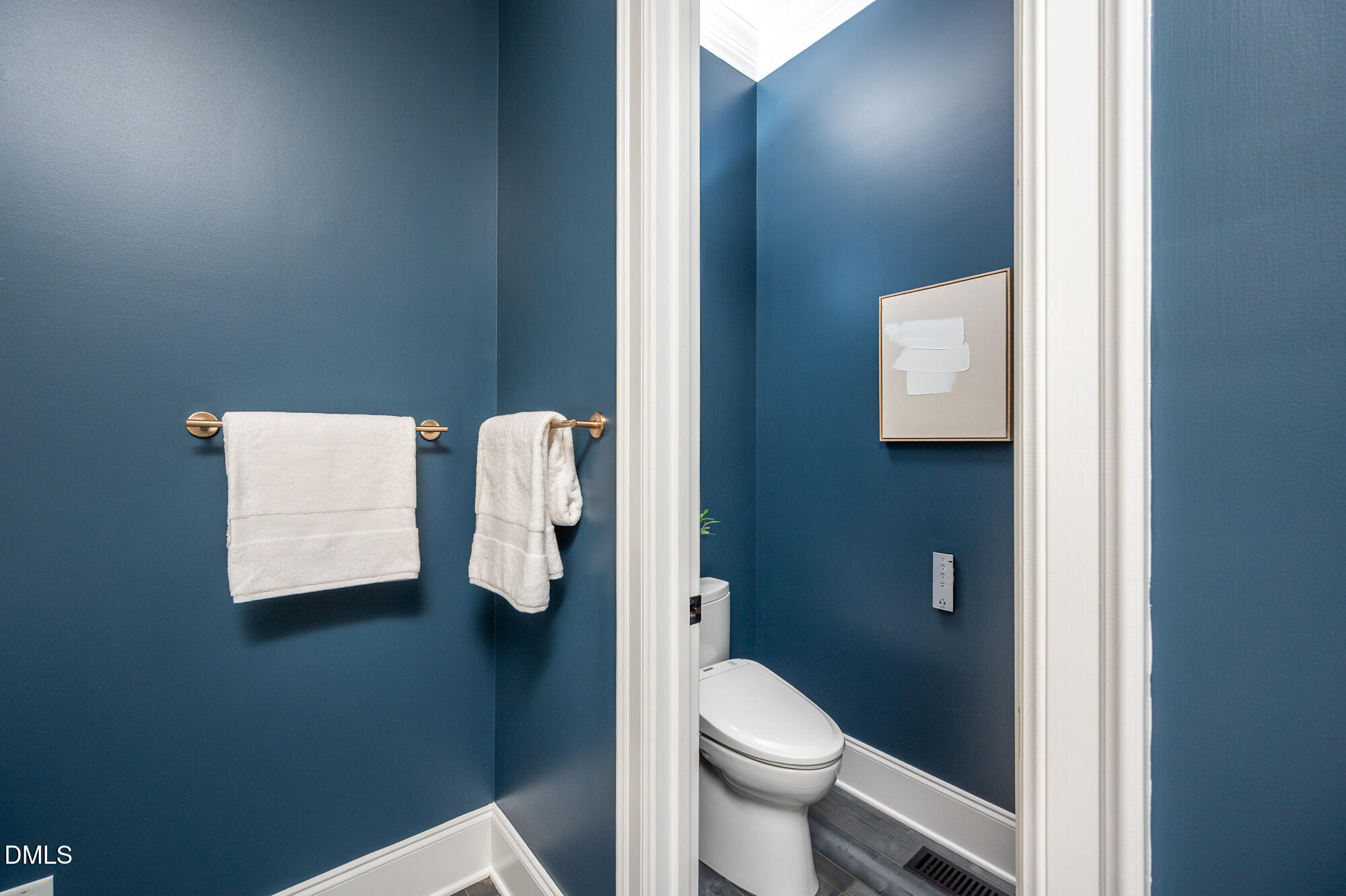 Bathroom with deep blue walls, white towels on brass bars, and a framed print beside the toilet.