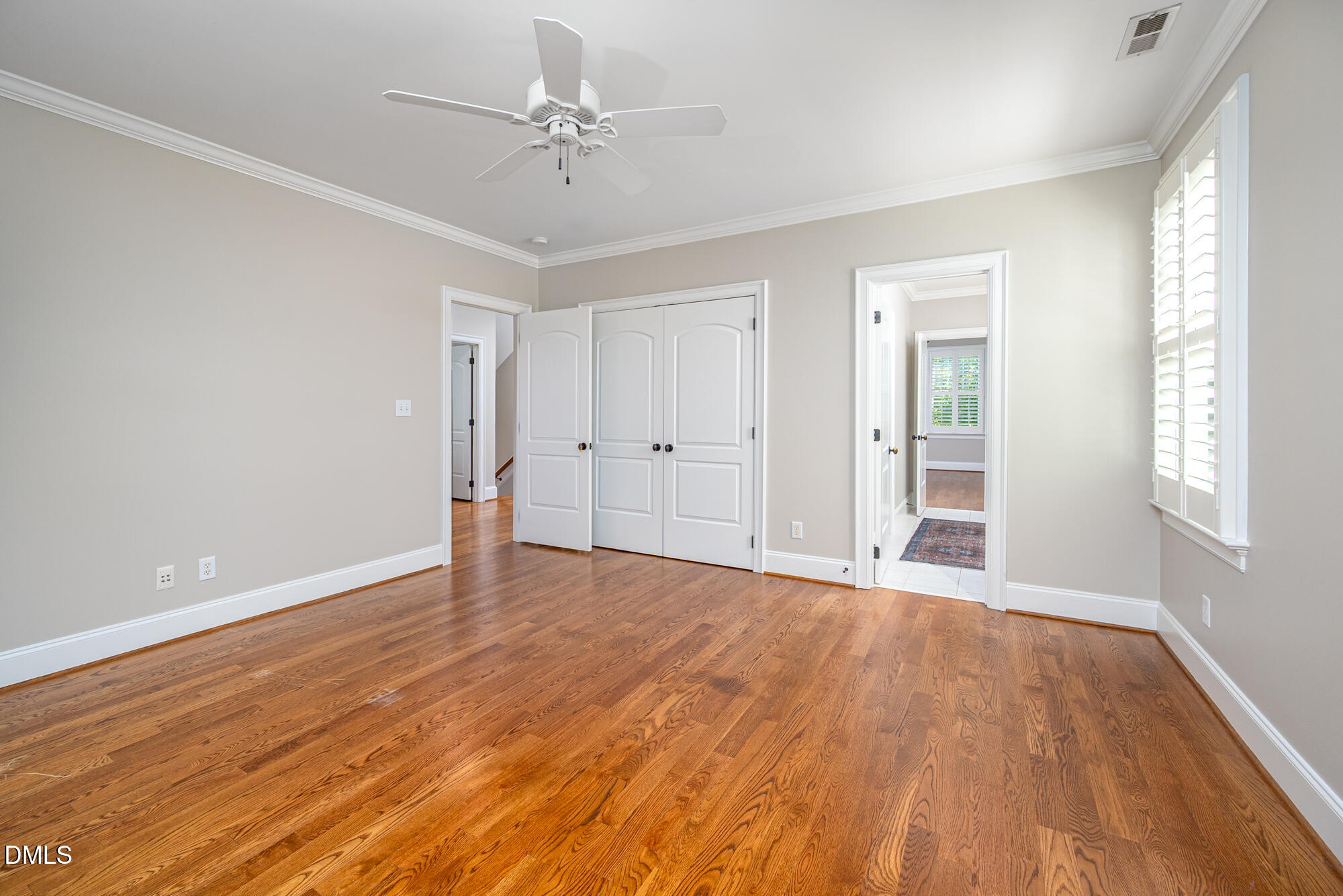 Empty bedroom with hardwood floors, double closet doors, a ceiling fan, and plantation shutters.