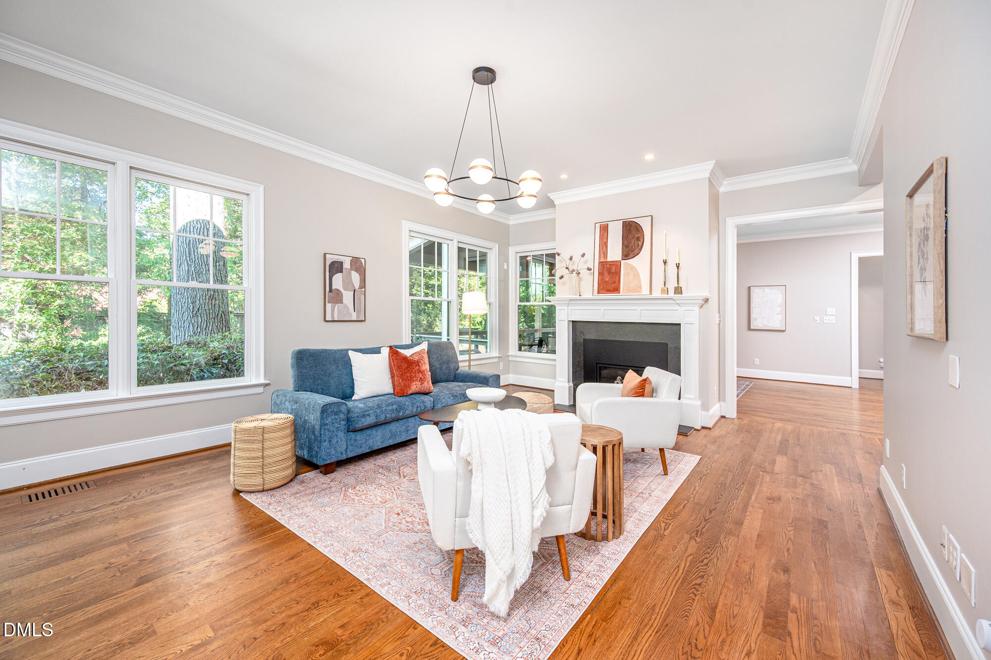 Staged living room with a fireplace, blue sofa, white chairs, and large windows overlooking trees.
