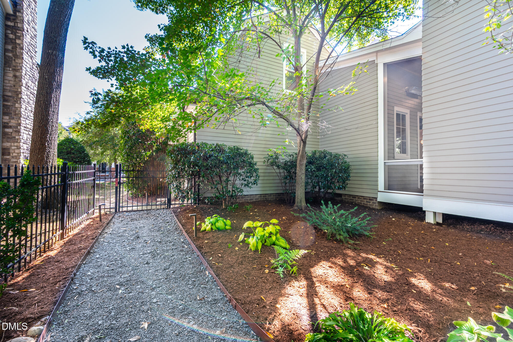 House side garden with a gravel path, black metal fence, and mature trees.