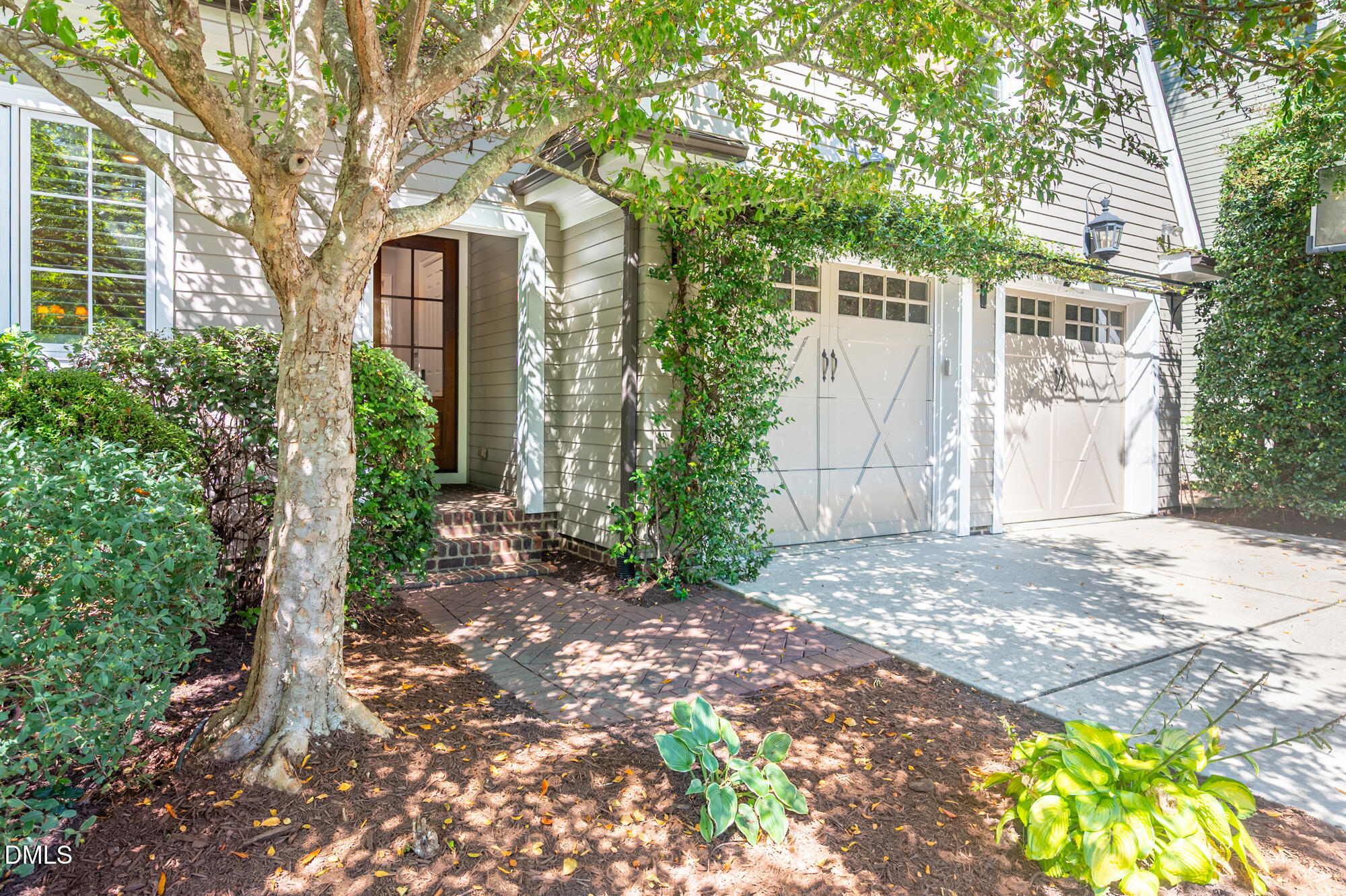 House entrance and two white garage doors framed by mature trees and landscaped shrubs.