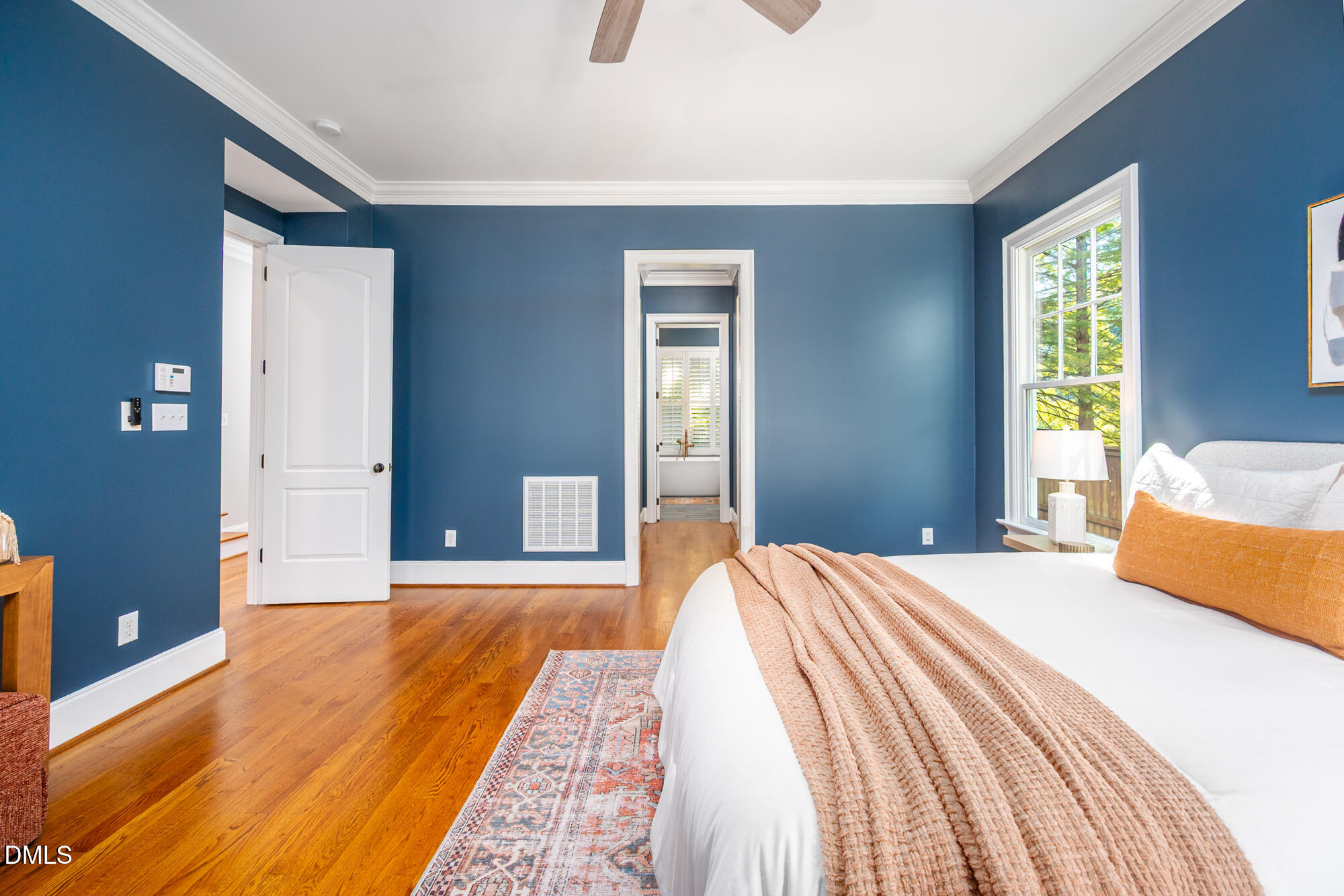 Blue bedroom with hardwood floors, a white bed, and a window overlooking leafy trees.