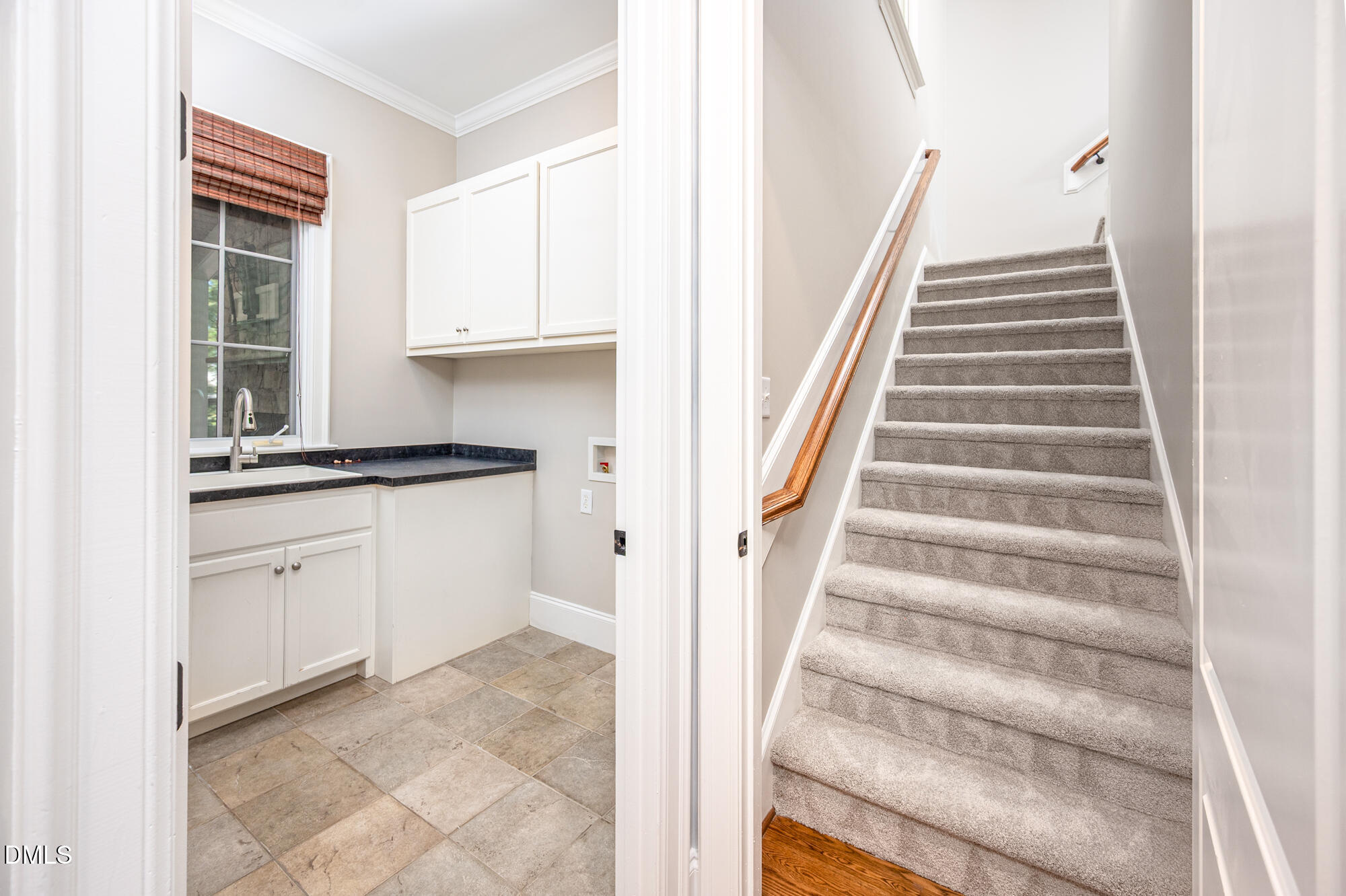 Tiled utility area with a sink and white cabinets beside a carpeted staircase with a wooden handrail.
