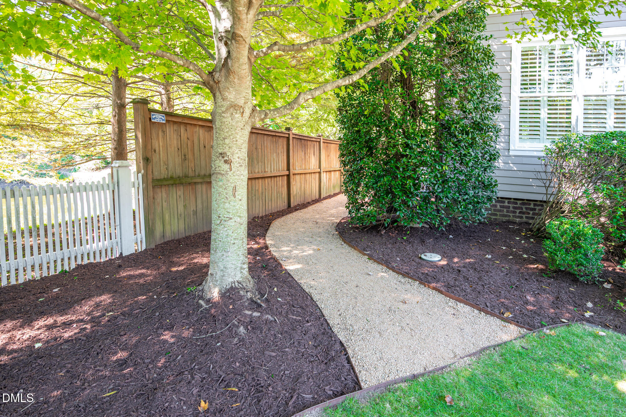 Curved gravel walkway beside a gray-sided house, bordered by a wooden fence, mature tree, and leafy shrubs.