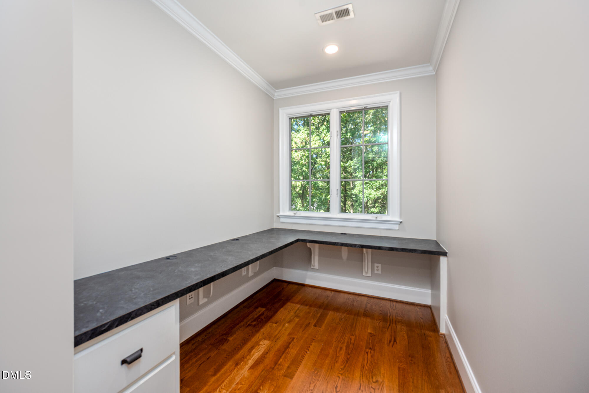 Empty home office with an L-shaped desk, hardwood floors, and a large window overlooking trees.