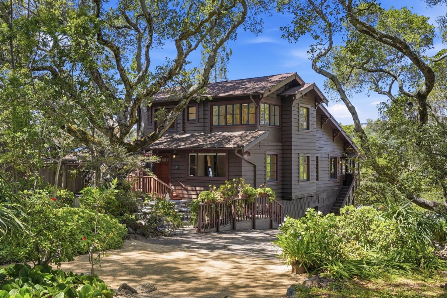 Two-story brown house among mature trees, with a landscaped garden, wooden deck, and stone-edged path.