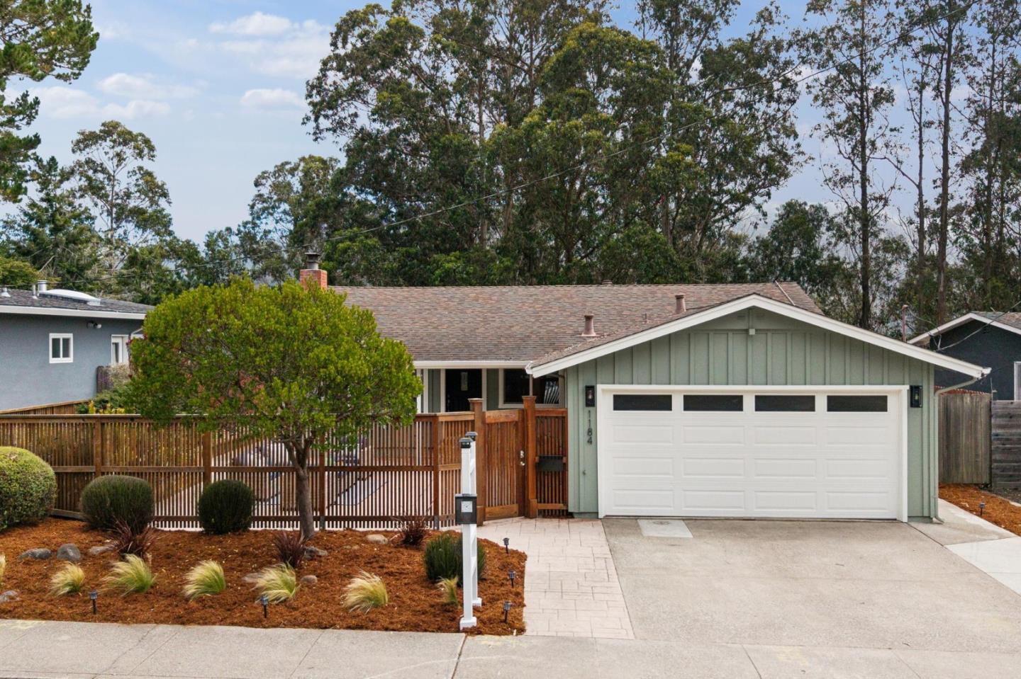 Sage-green single-story home with a wide white garage door, wooden fence, and landscaped front yard.