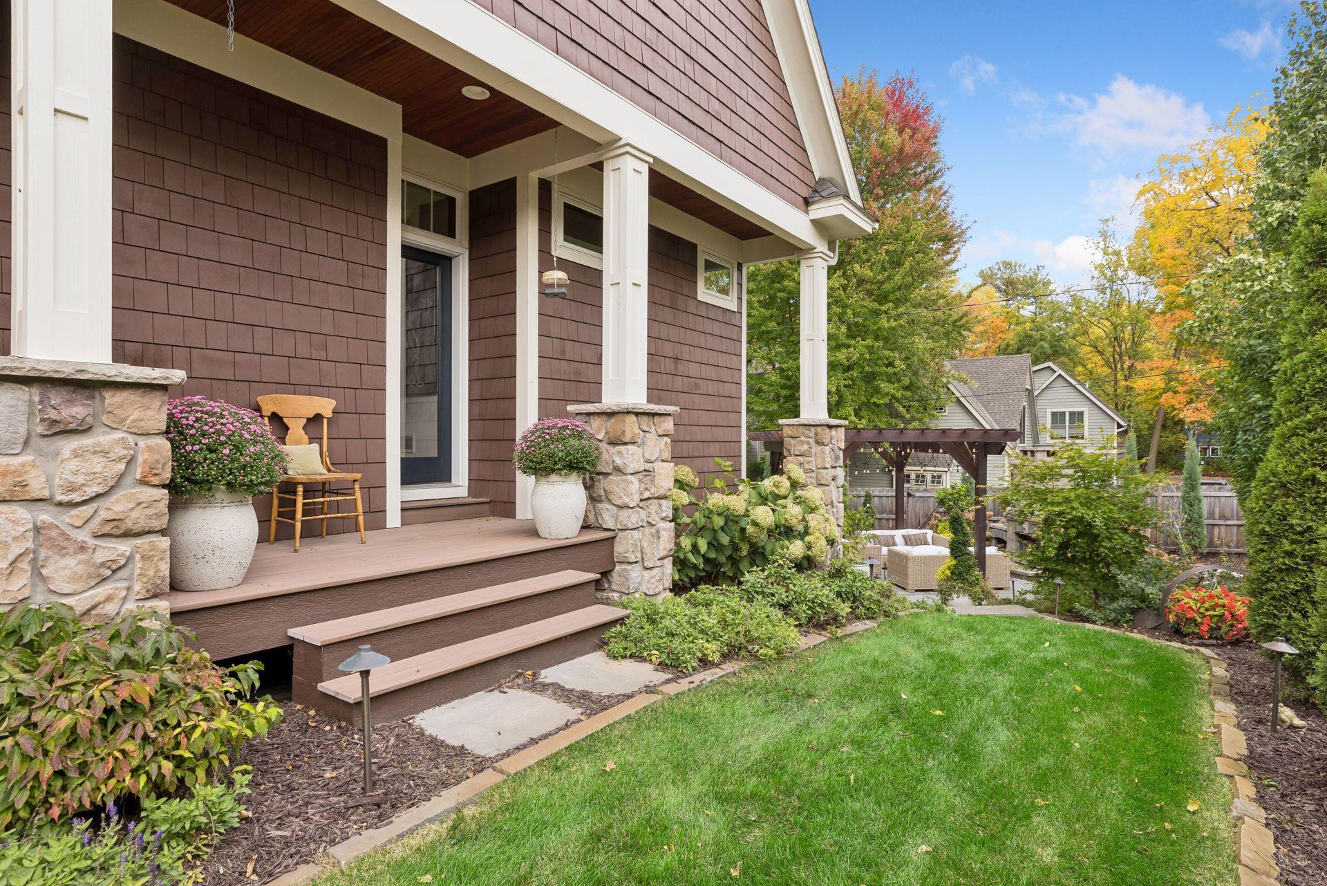 Brown-shingled home with a stone-columned porch, flowering planters, and a landscaped backyard patio.