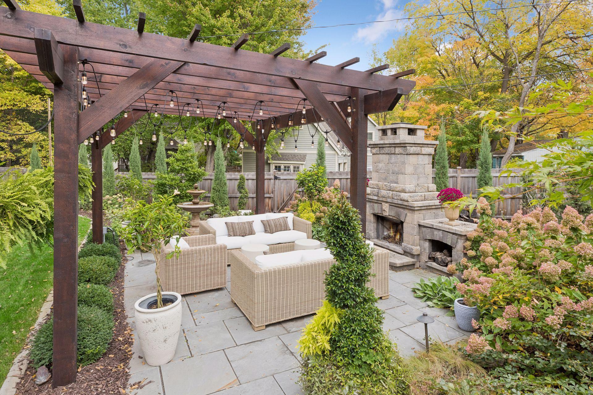 Patio beneath a wooden pergola, with wicker seating and a stone fireplace amid autumn landscaping.