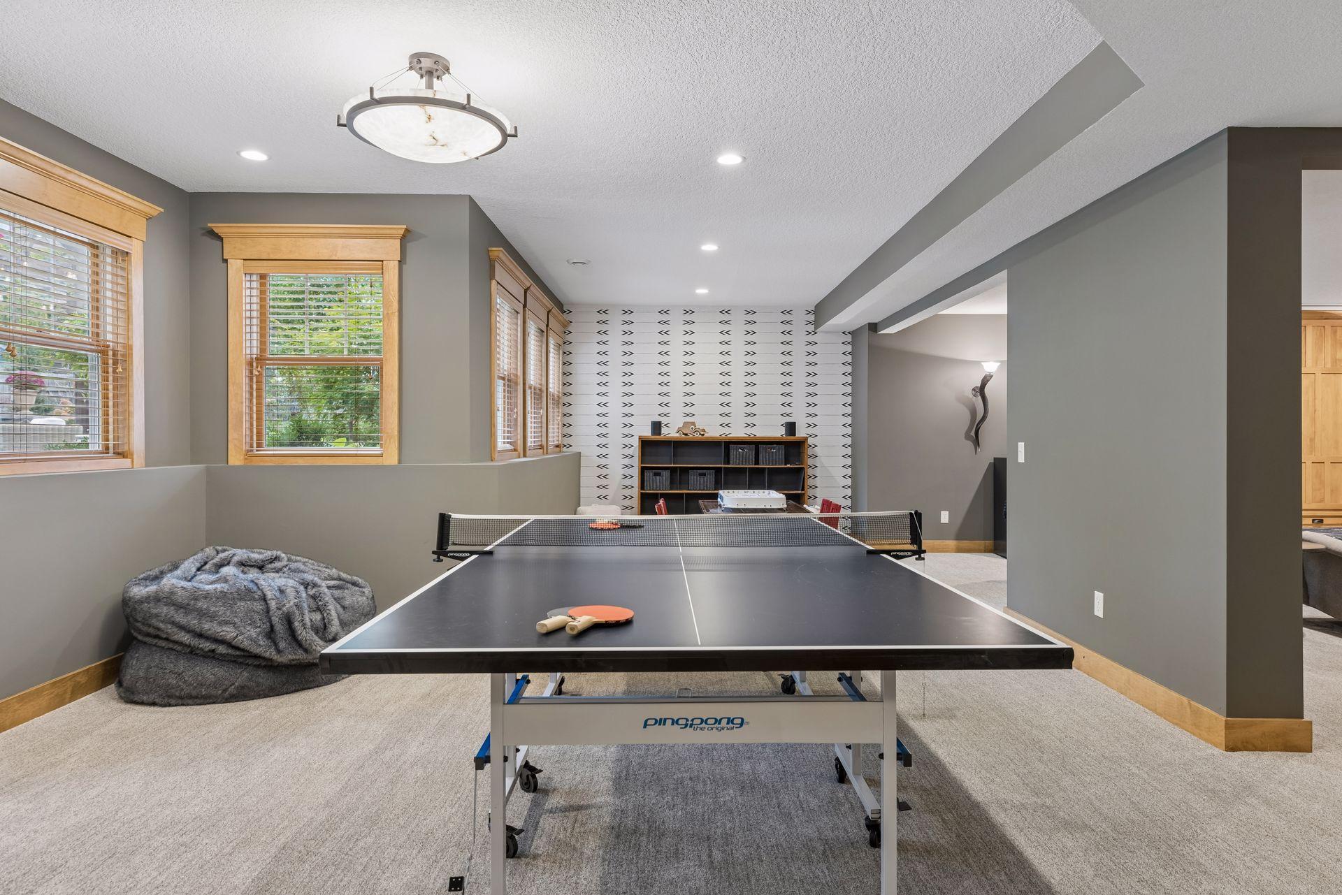 Furnished recreation room with a ping-pong table, patterned accent wall, and large wood-trimmed windows.