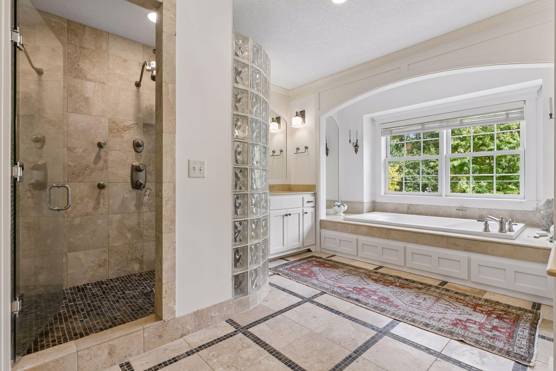Bathroom with a tiled walk-in shower, glass-block divider, and built-in tub beneath a large window.