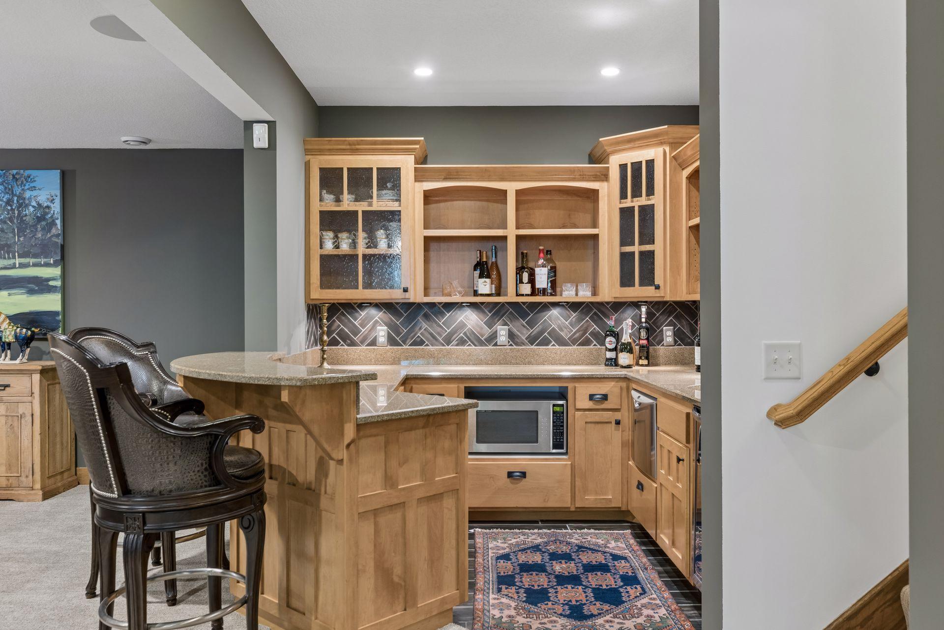 Wet bar with light wood cabinetry, herringbone tile backsplash, granite counters, and upholstered bar stools.