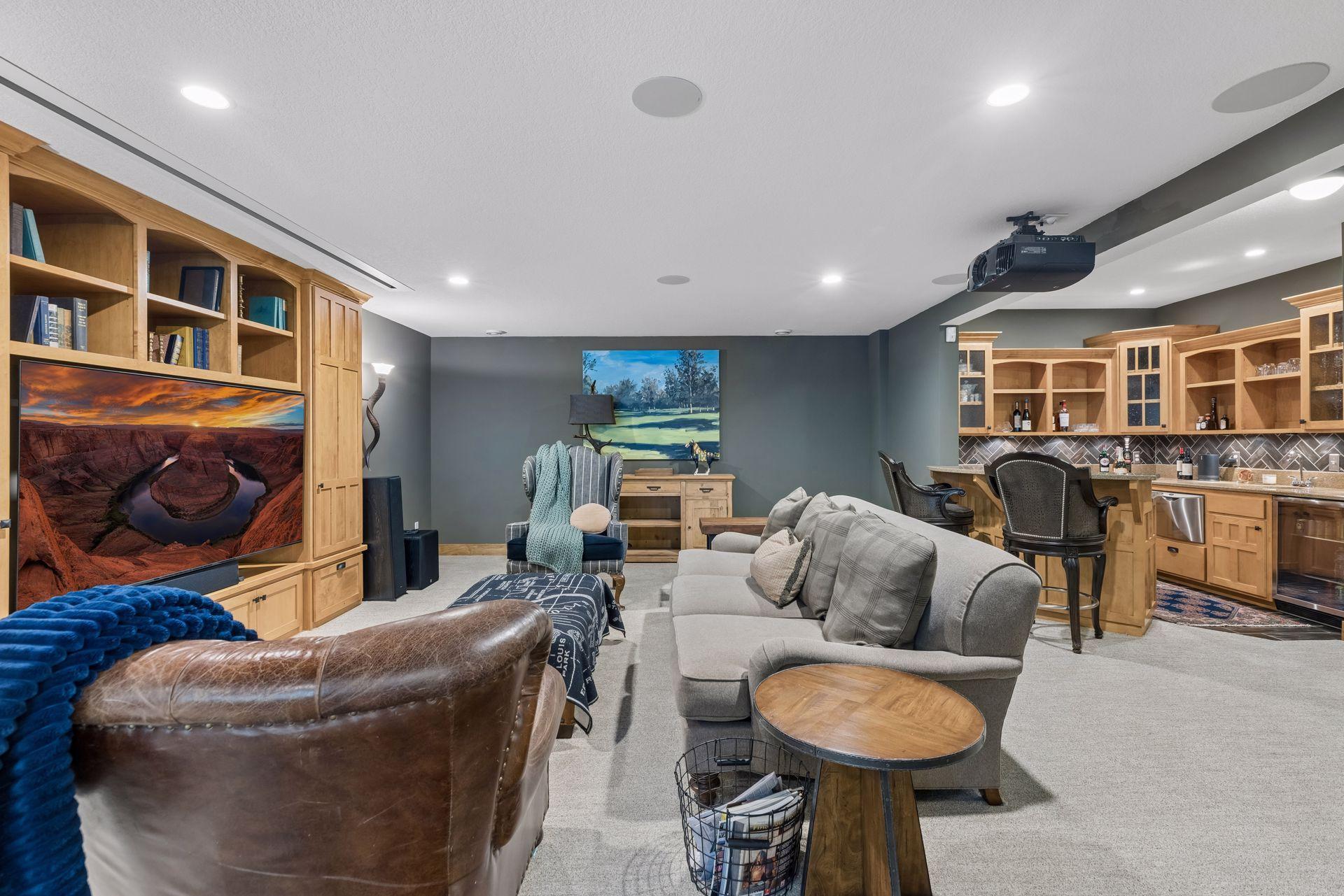 Basement media room with custom wood cabinetry, a ceiling-mounted projector, gray seating, and a wet bar.