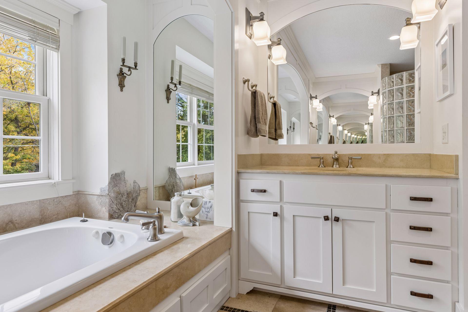 Bright bathroom with a soaking tub beside tall windows and a long mirrored vanity.