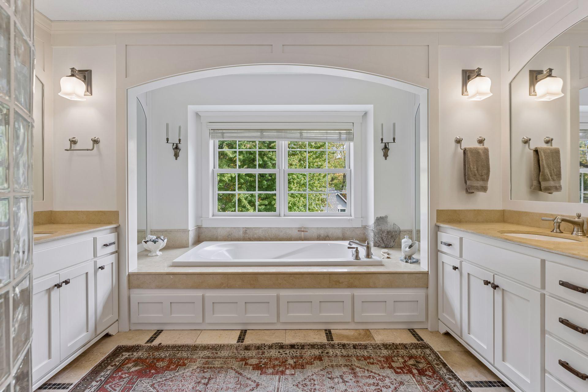 Bathroom with a soaking tub beneath a large window, framed by an arched alcove and flanked by dual vanities.