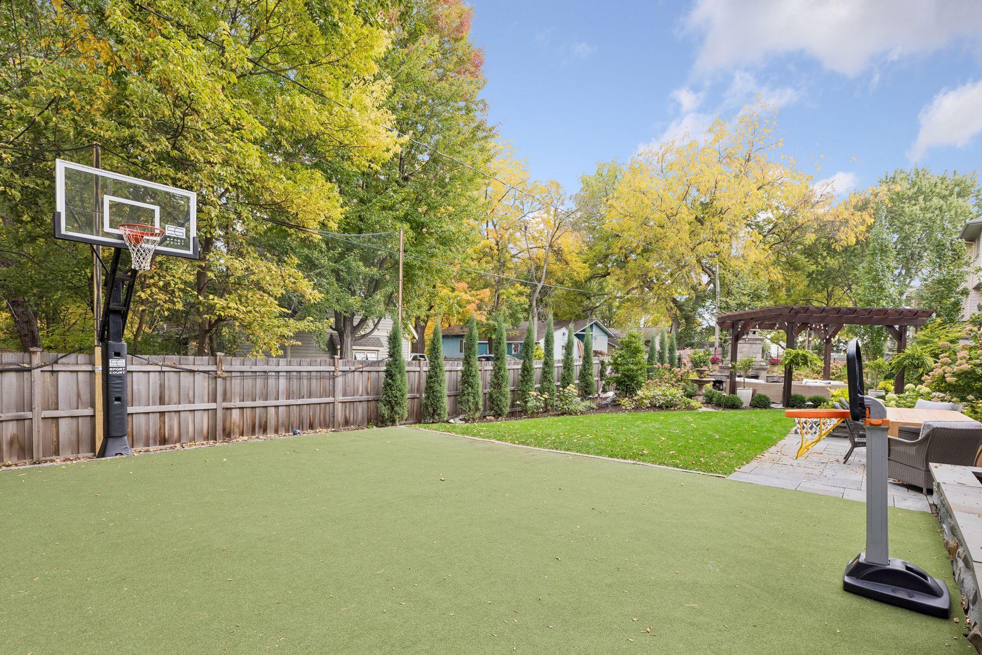 Backyard basketball court with green turf beside a pergola-covered patio and autumn trees.