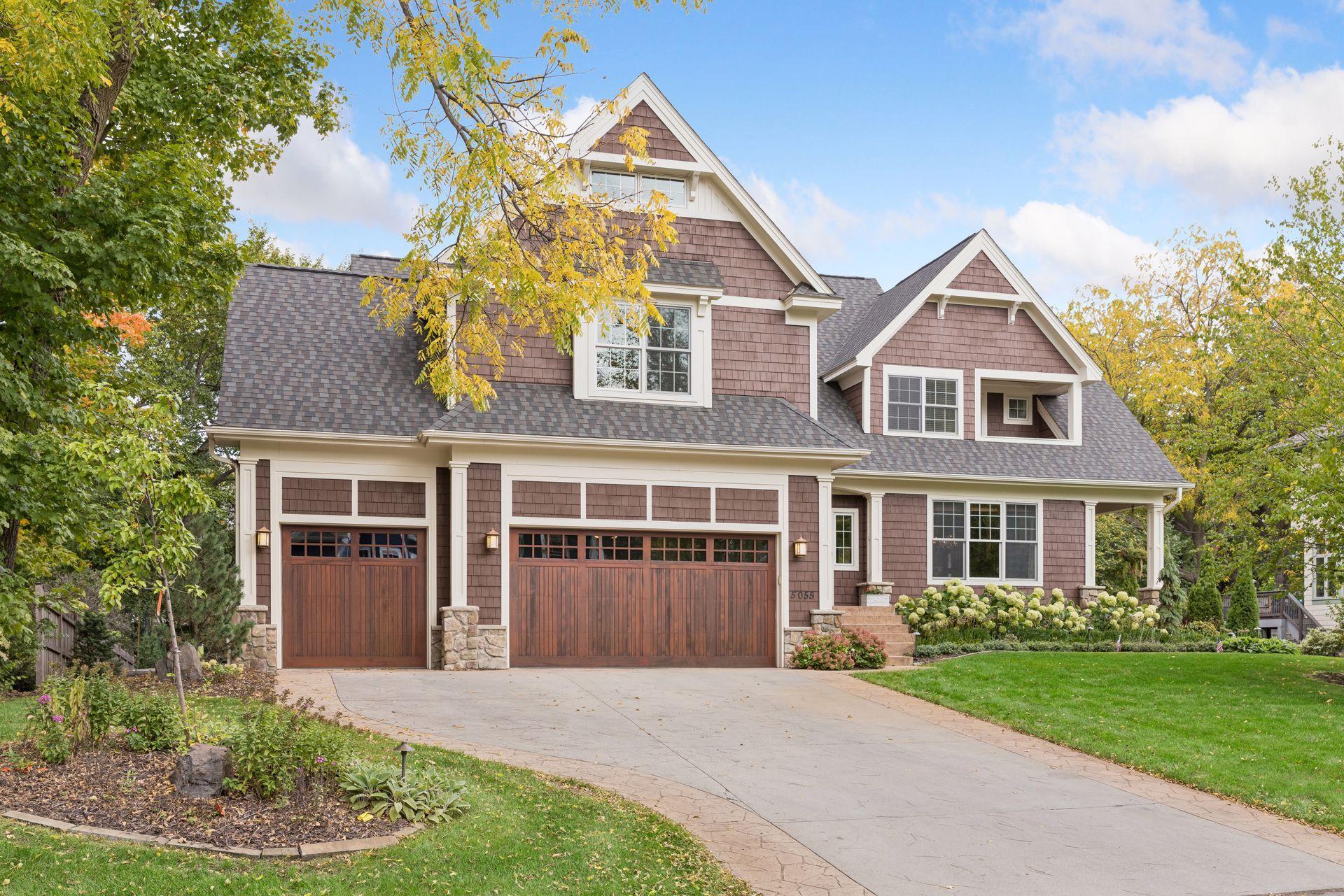 Two-story brown-shingled home with white-trimmed gables, wooden garage doors, and autumn trees.