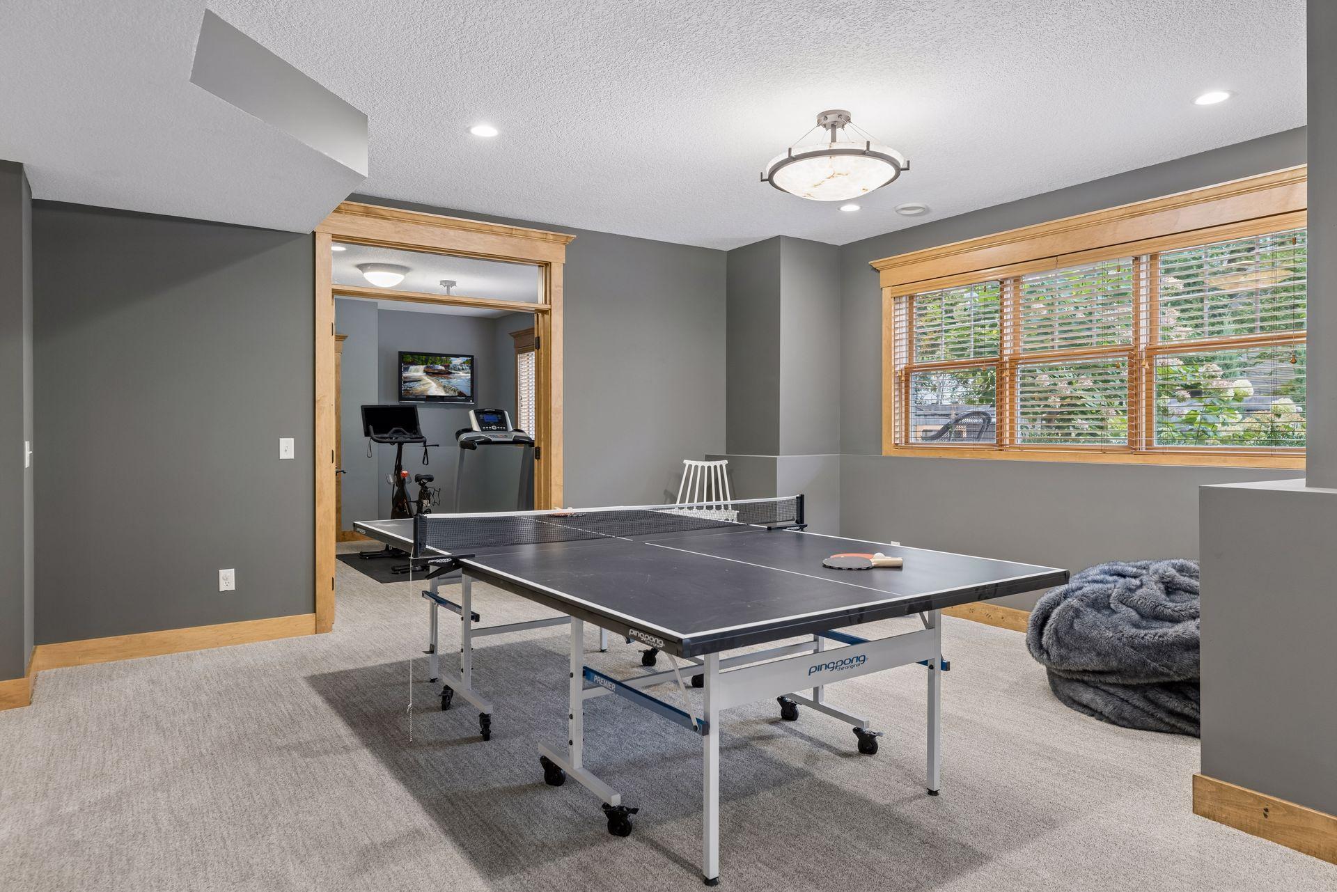 Recreation room with a ping-pong table, exercise equipment, and a wide window with wooden blinds.