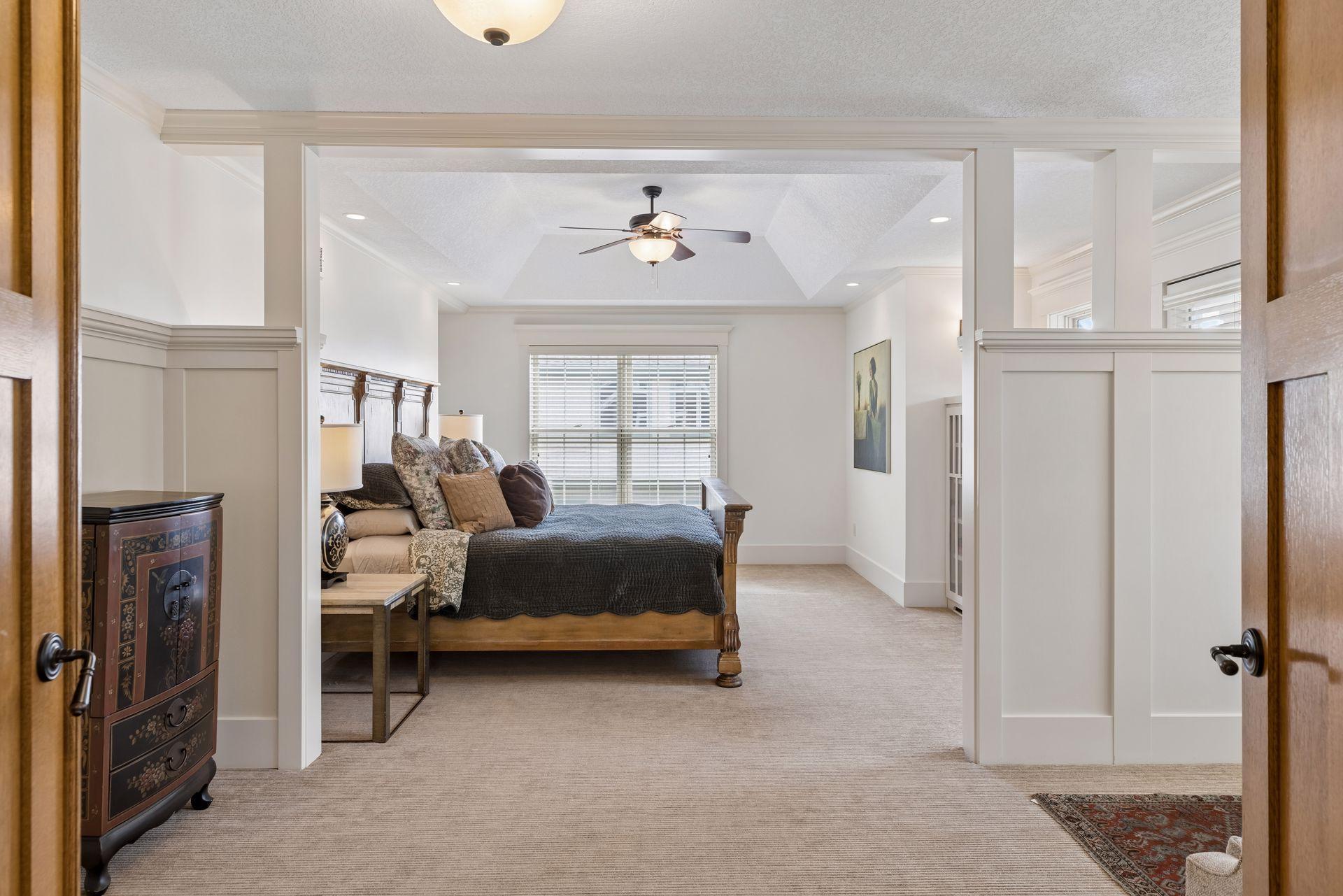 Furnished bedroom with a wooden bed, tray ceiling and ceiling fan, viewed through open double doors.