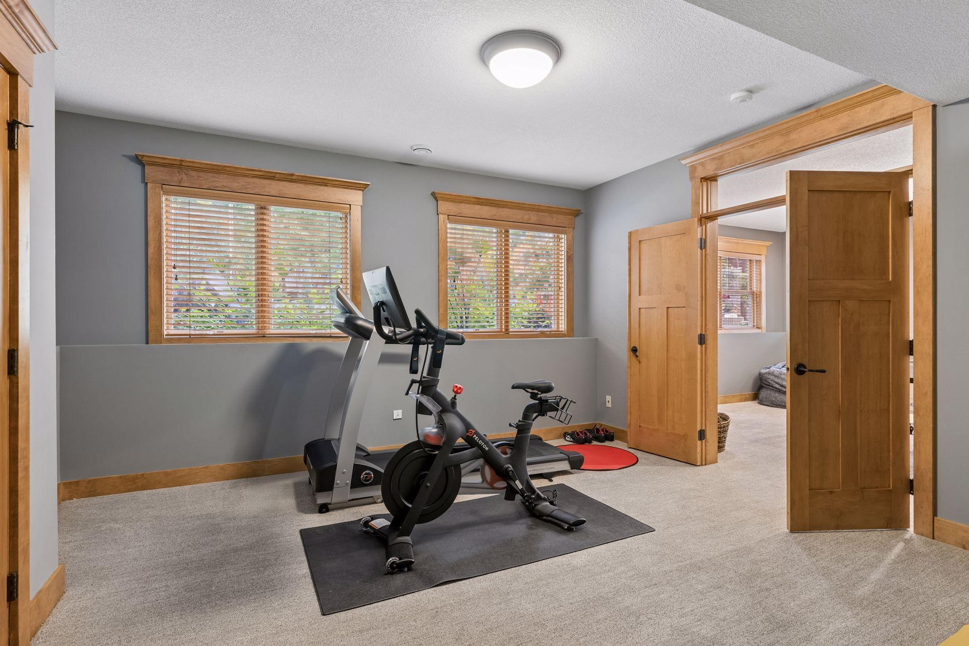 Home gym with a stationary bike and treadmill beneath wood-trimmed windows.