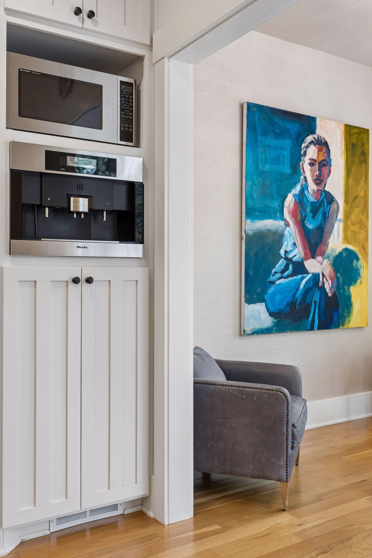 Built-in microwave and coffee maker beside a gray armchair and colorful portrait painting.