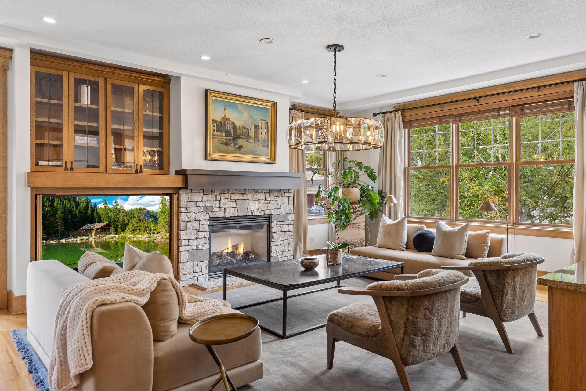 Living room with a stone fireplace, wood cabinetry, and large windows overlooking trees.