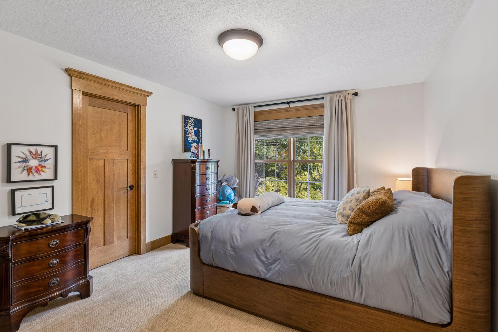 Bedroom with a large wooden bed, gray bedding, and a window overlooking leafy trees.