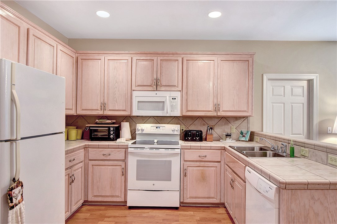 Kitchen with light wood cabinets, white appliances, tiled counters, and a double sink.