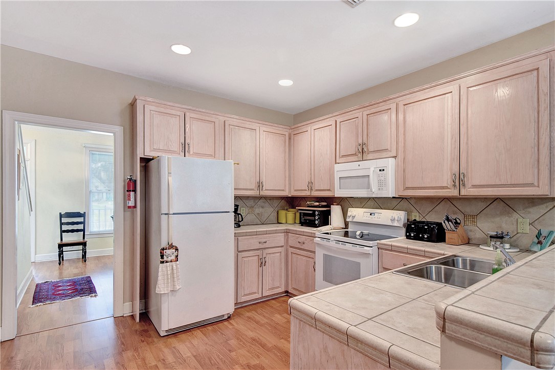 Kitchen with pale wood cabinetry, tiled counters, white appliances, and a peninsula sink.