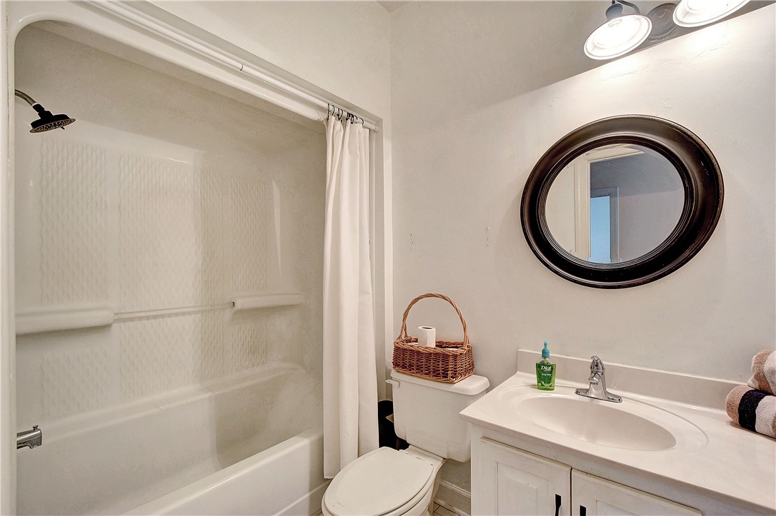 Bathroom with a white tub and shower, wicker basket on the toilet, and oval mirror above the vanity.