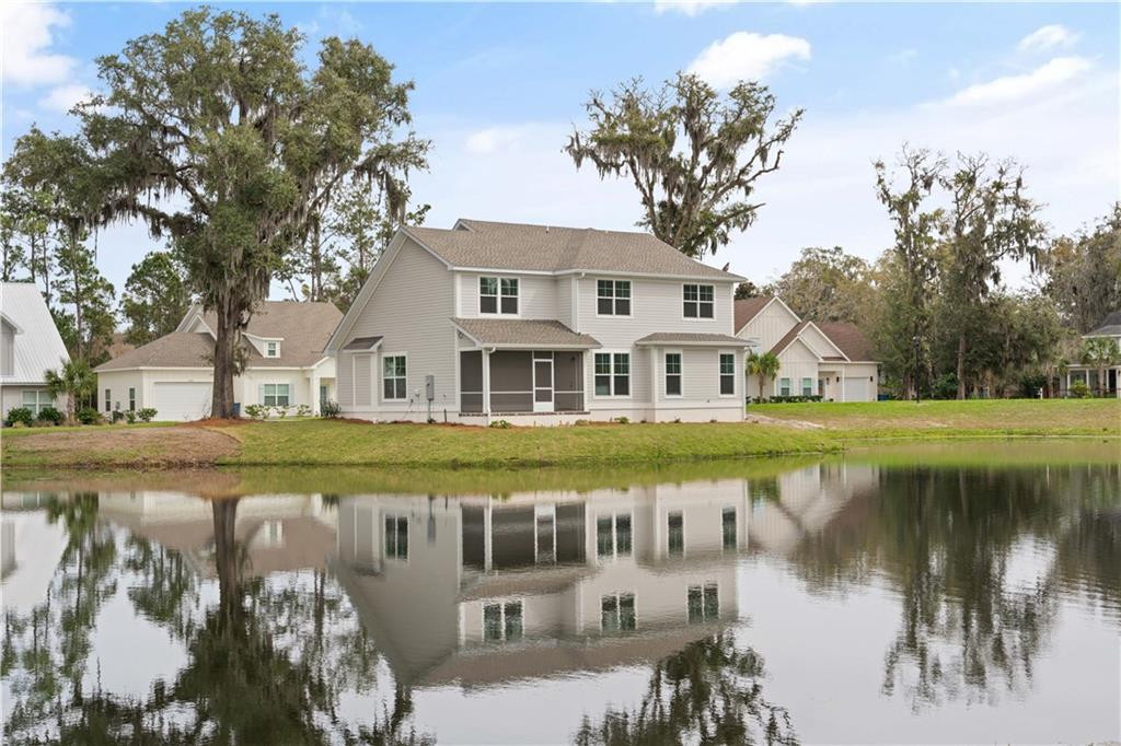 Two-story house with a covered porch reflected in a pond, framed by mature trees.