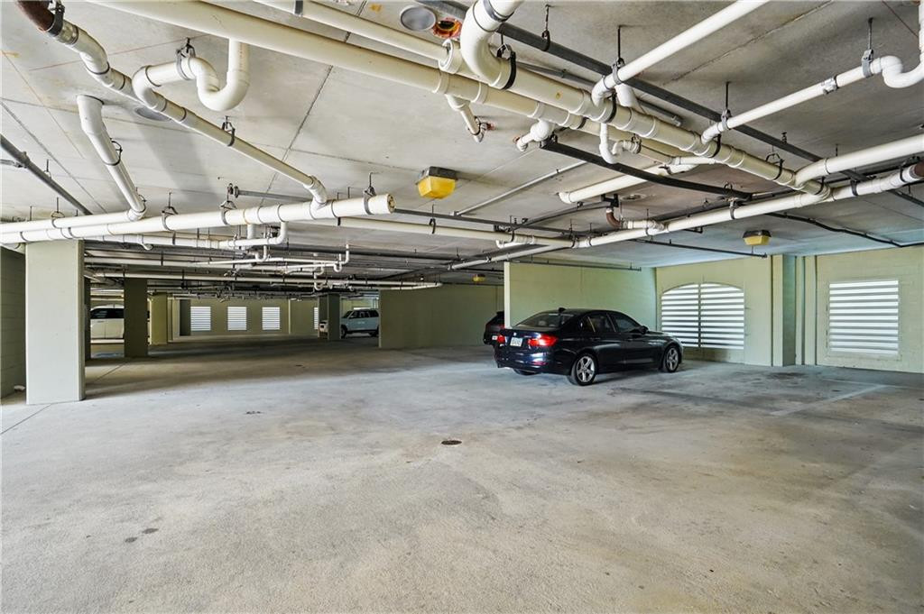 Concrete parking garage with exposed pipes overhead and cars parked beside louvered openings.