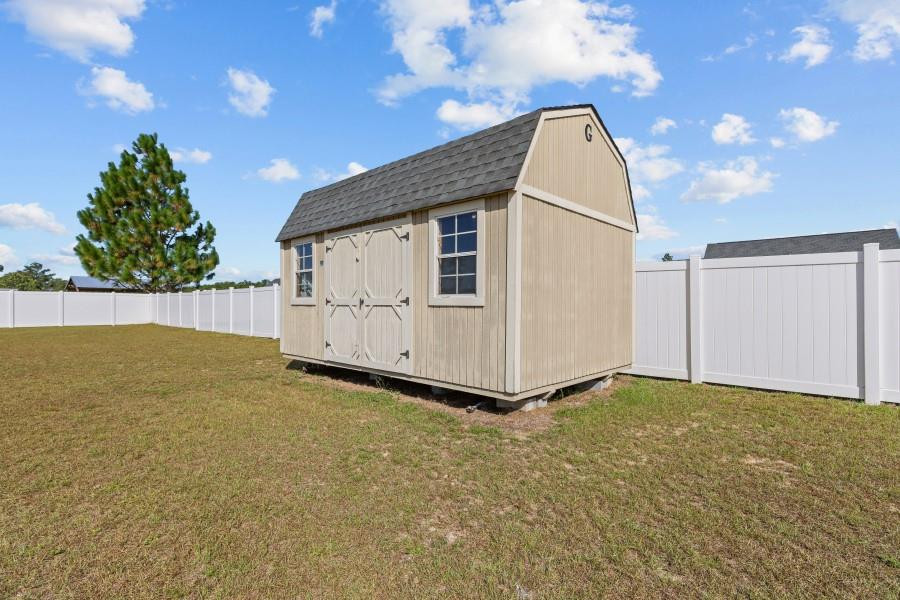 Beige storage shed with a gambrel roof, double barn doors, and two windows inside a white fence.