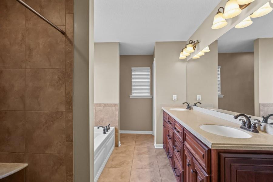 Bathroom with a long double-sink vanity, reddish wood cabinets, and tan tile flooring.